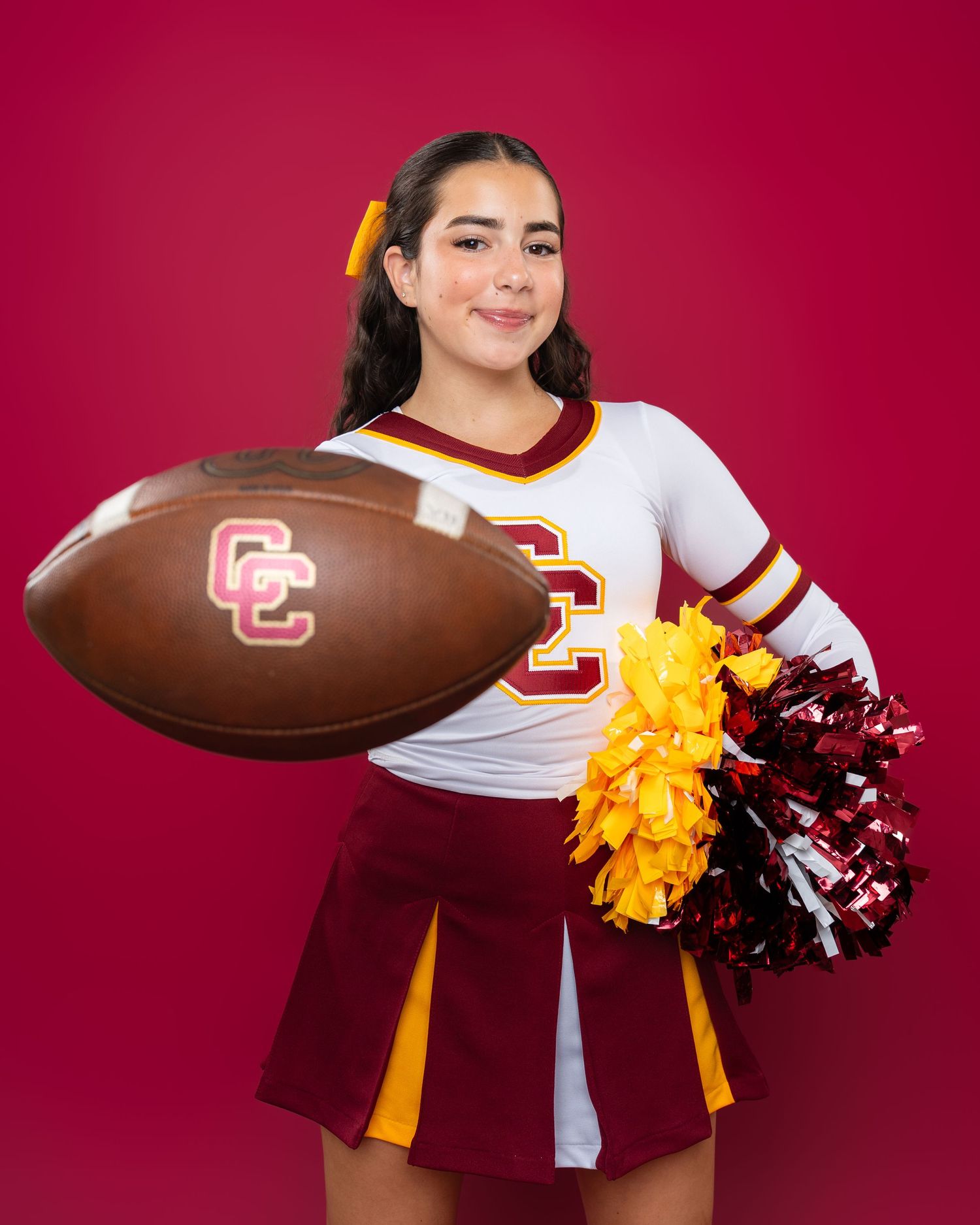 Basketball cheerleader poses with football and pom poms against red background.