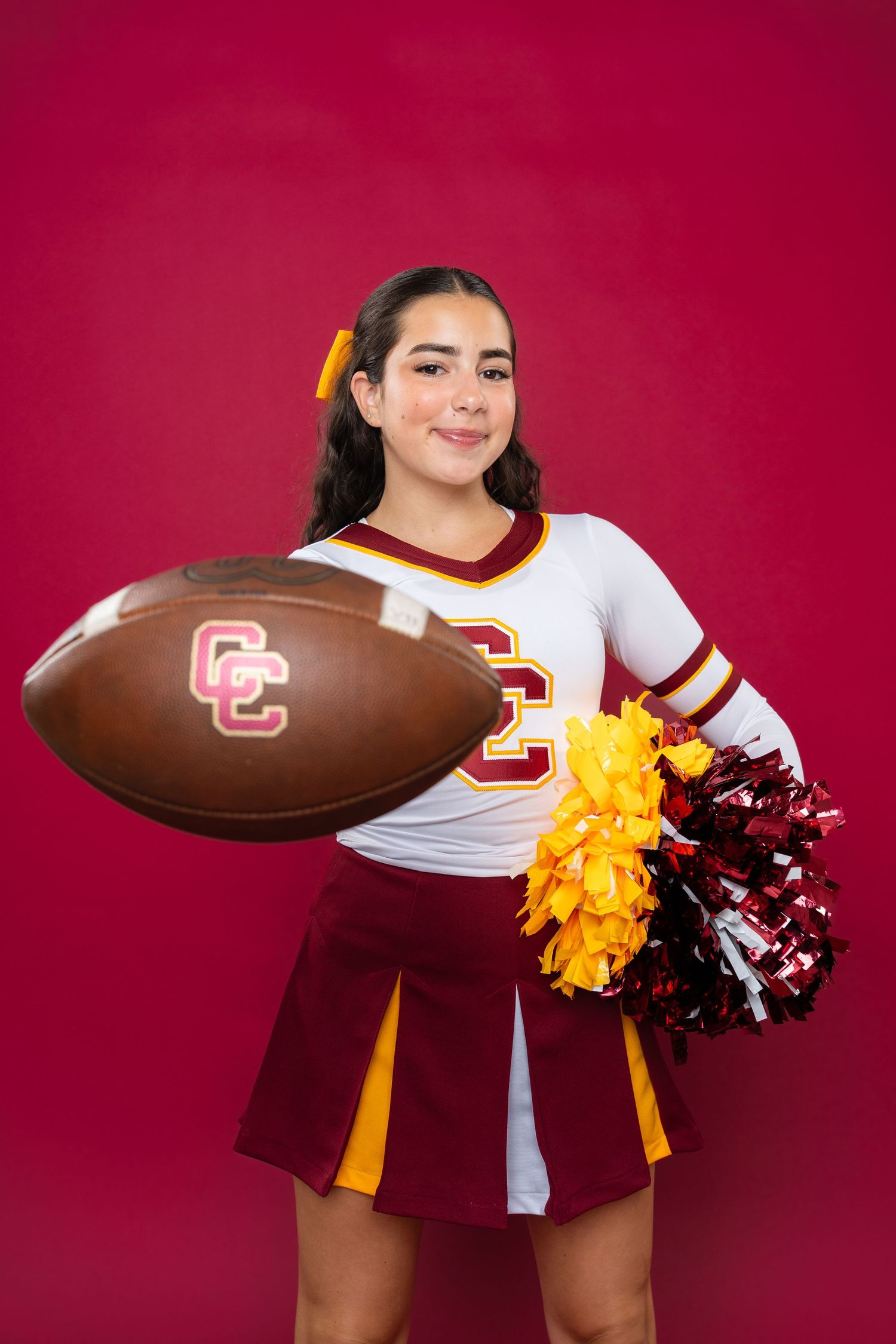 College cheerleader in white and burgundy uniform poses with football and pom poms against red background.