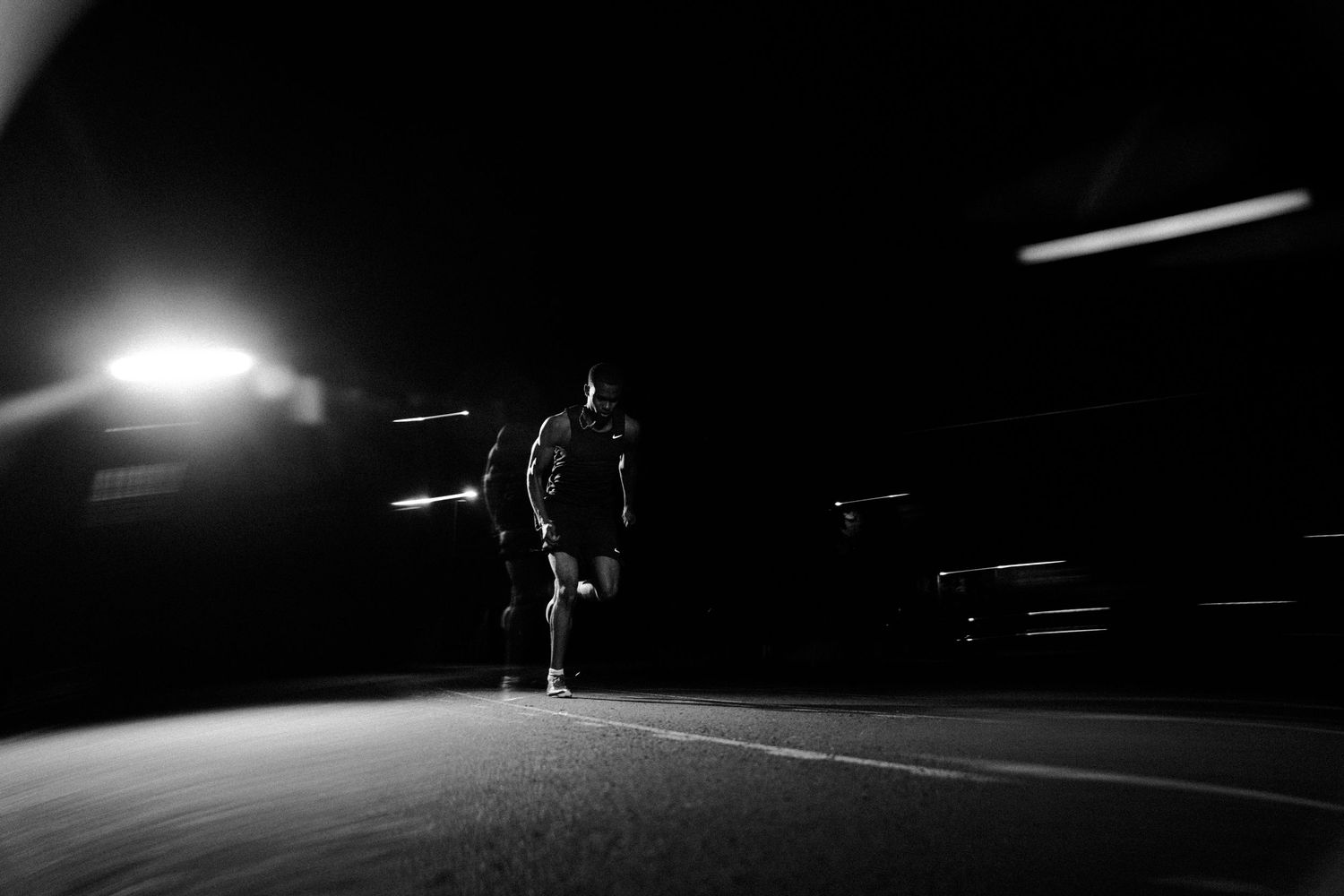 A lone figure stands on a dark street illuminated by bright headlights in a dramatic black and white photo.