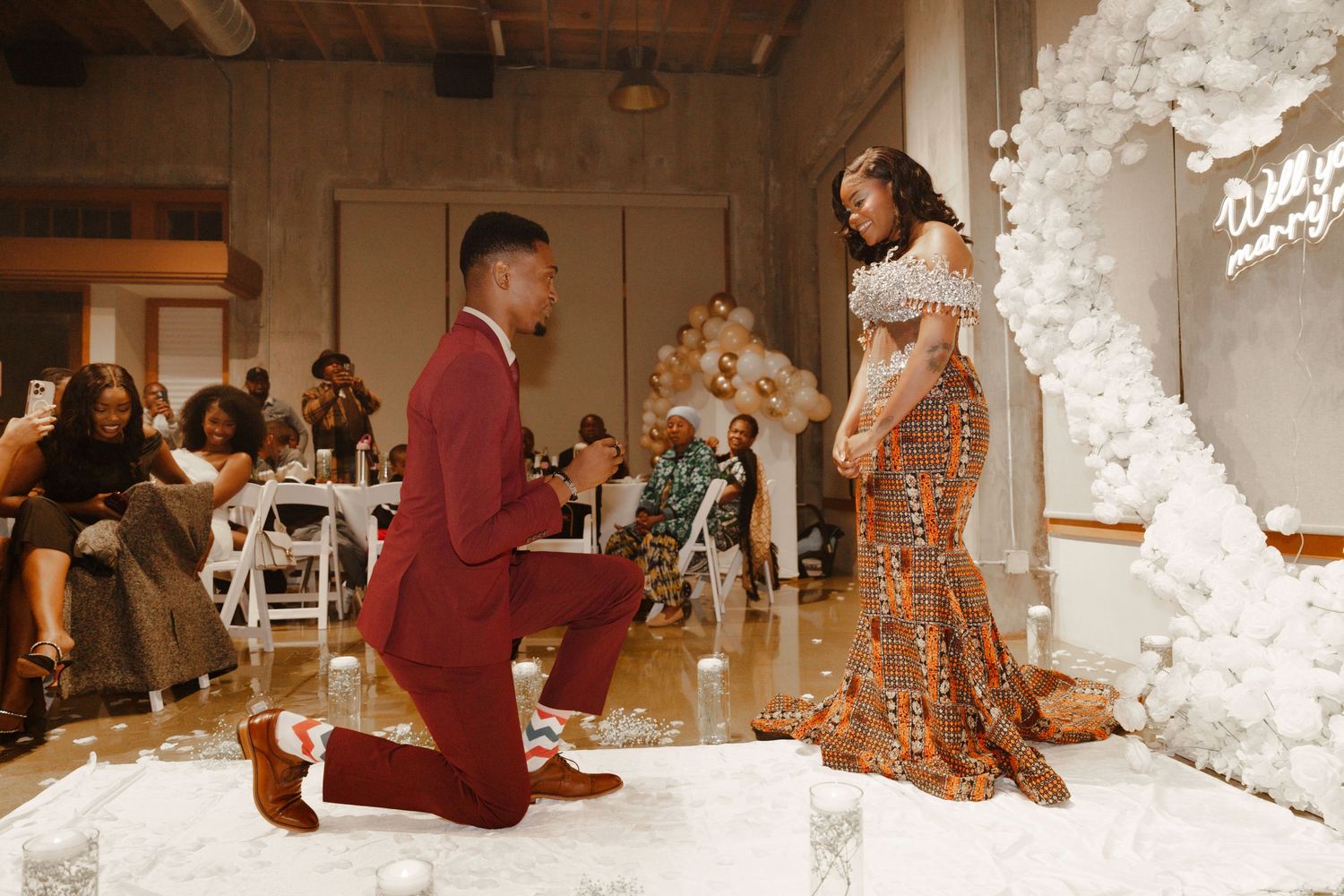 Man in red suit proposes on one knee to woman in elegant gown at romantic indoor proposal event.