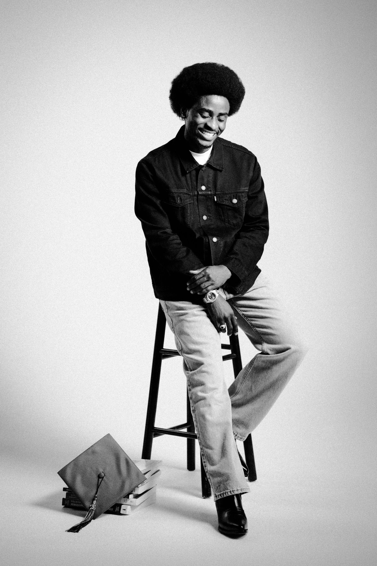 Black and white photo of a smiling man with an afro, sitting on a stool in a denim jacket and jeans.