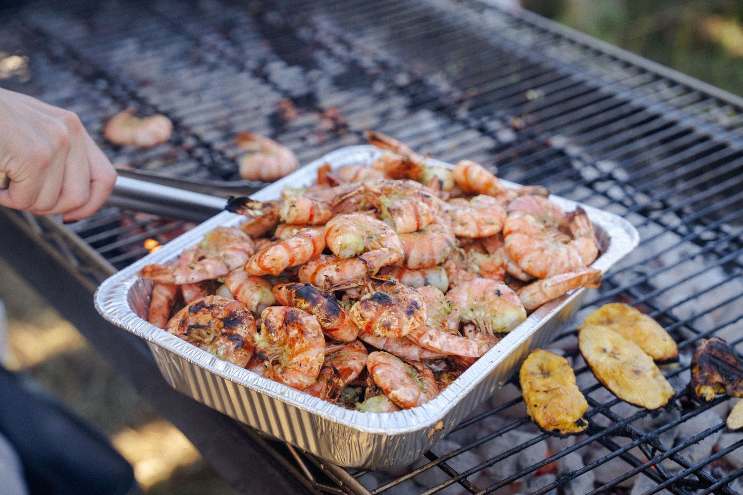 Grilled shrimp in aluminum foil pan on BBQ grill with tongs, perfect for outdoor seafood cookout.