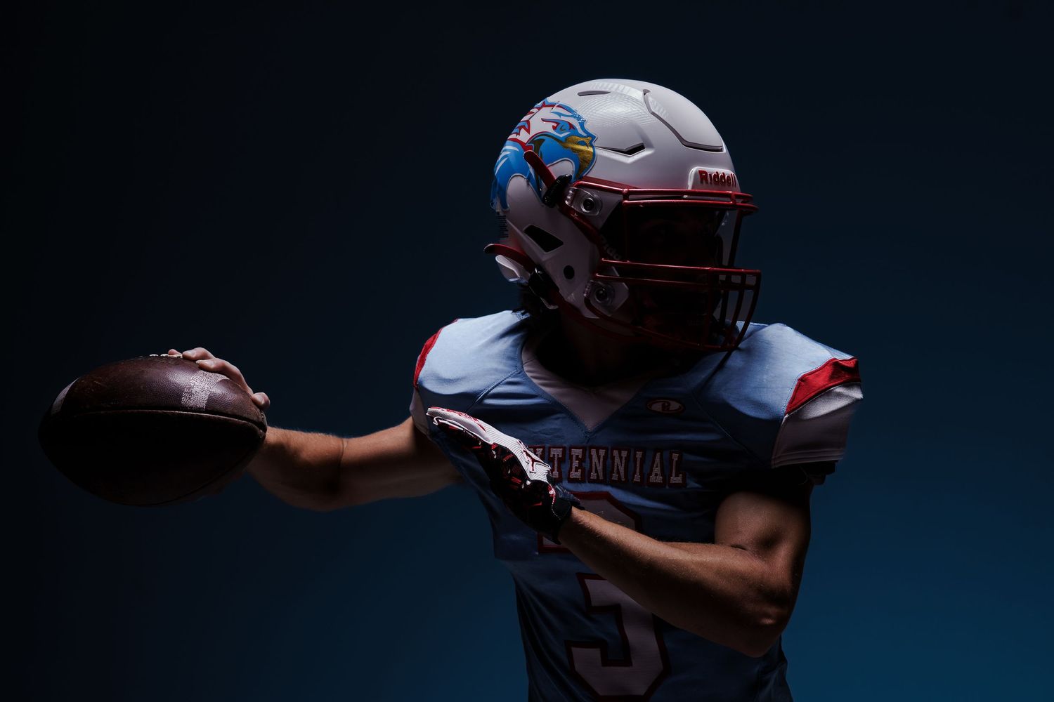 Football player in protective gear holding a ball against dark background.