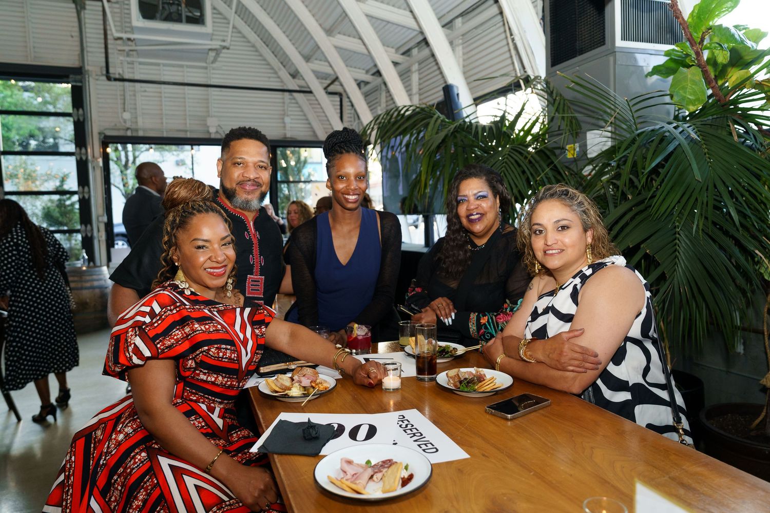 Five friends smile together at a restaurant table with food and drinks during a social gathering.