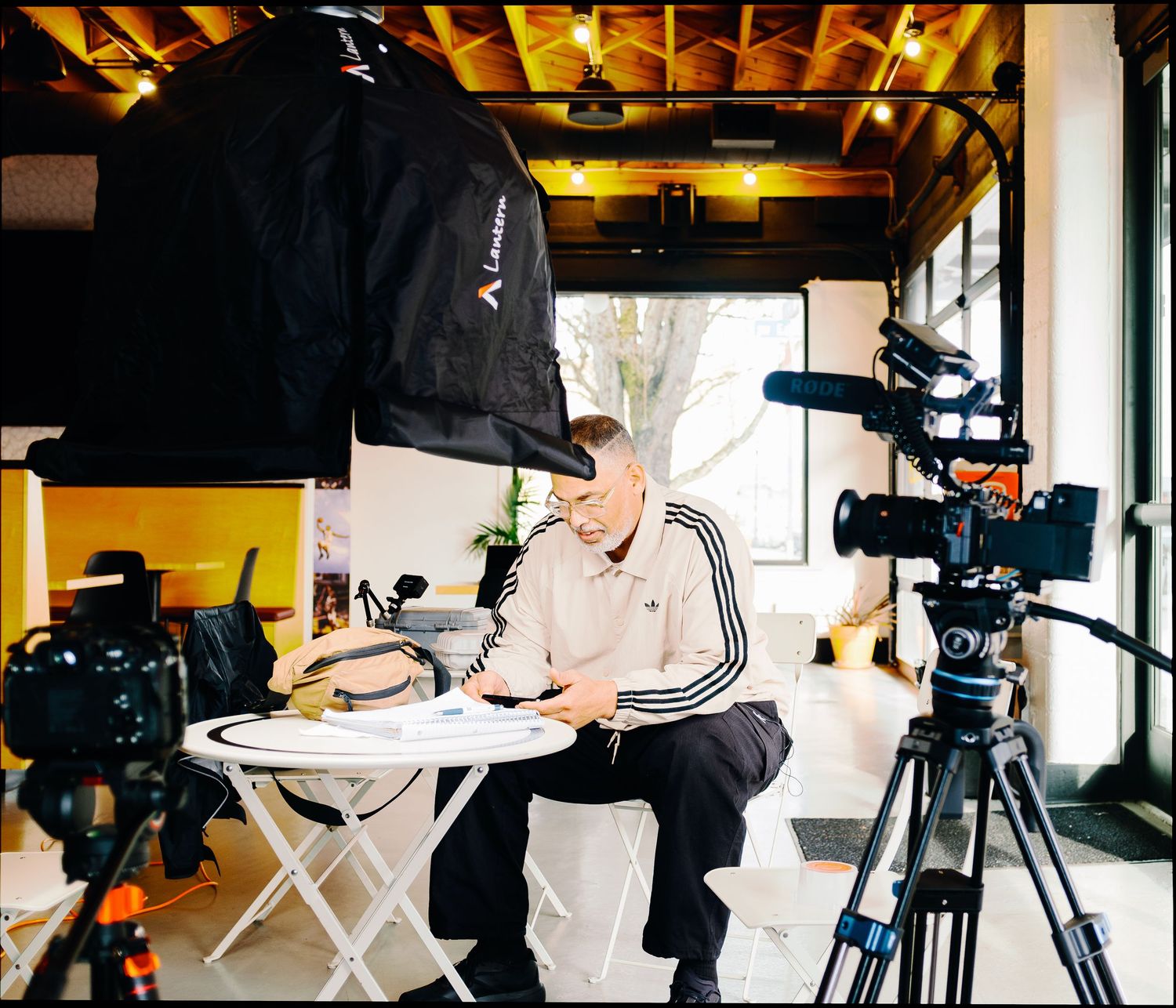 Man in Adidas tracksuit reviewing documents on a folding table surrounded by professional cameras and studio lighting equipment.