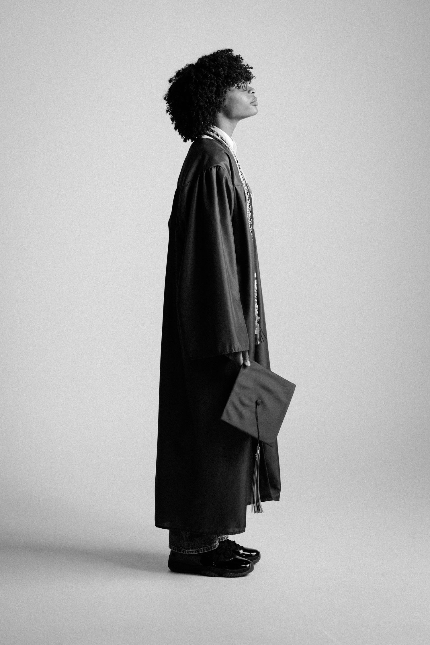 Black and white photo of a graduate in cap and gown holding mortarboard, standing sideways against light background.