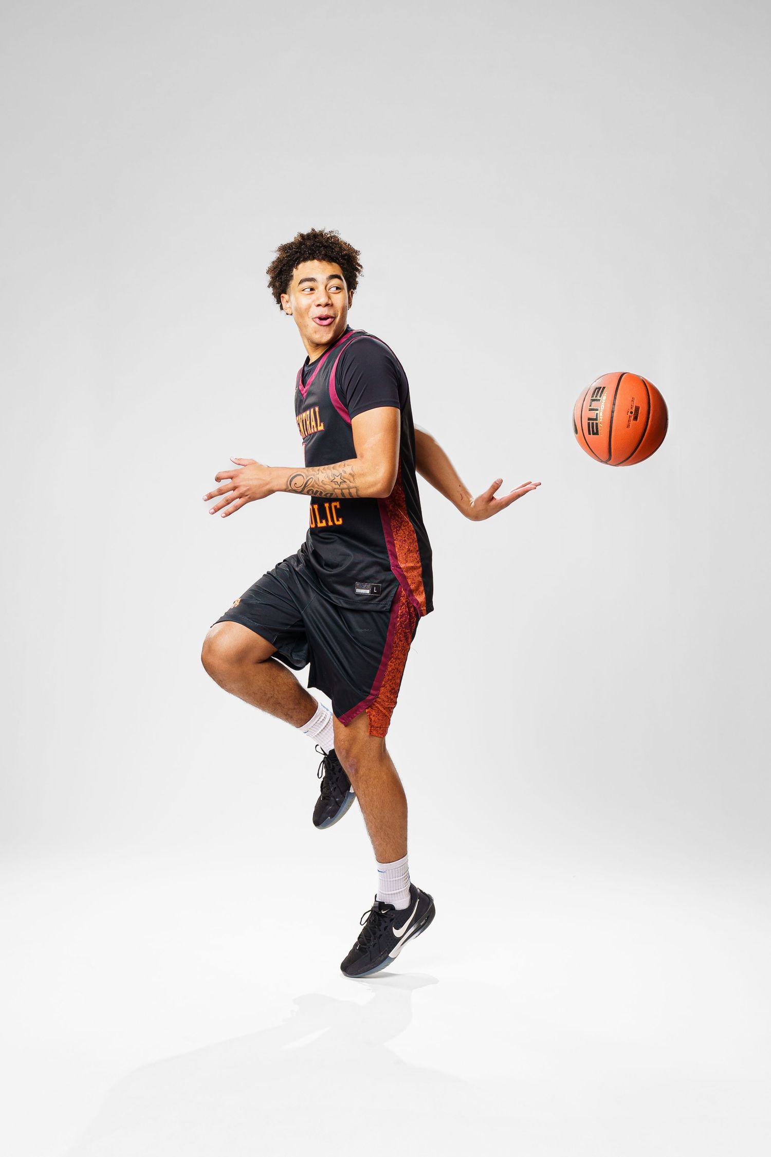 Basketball player in black uniform jumps and reaches for orange ball against white background.