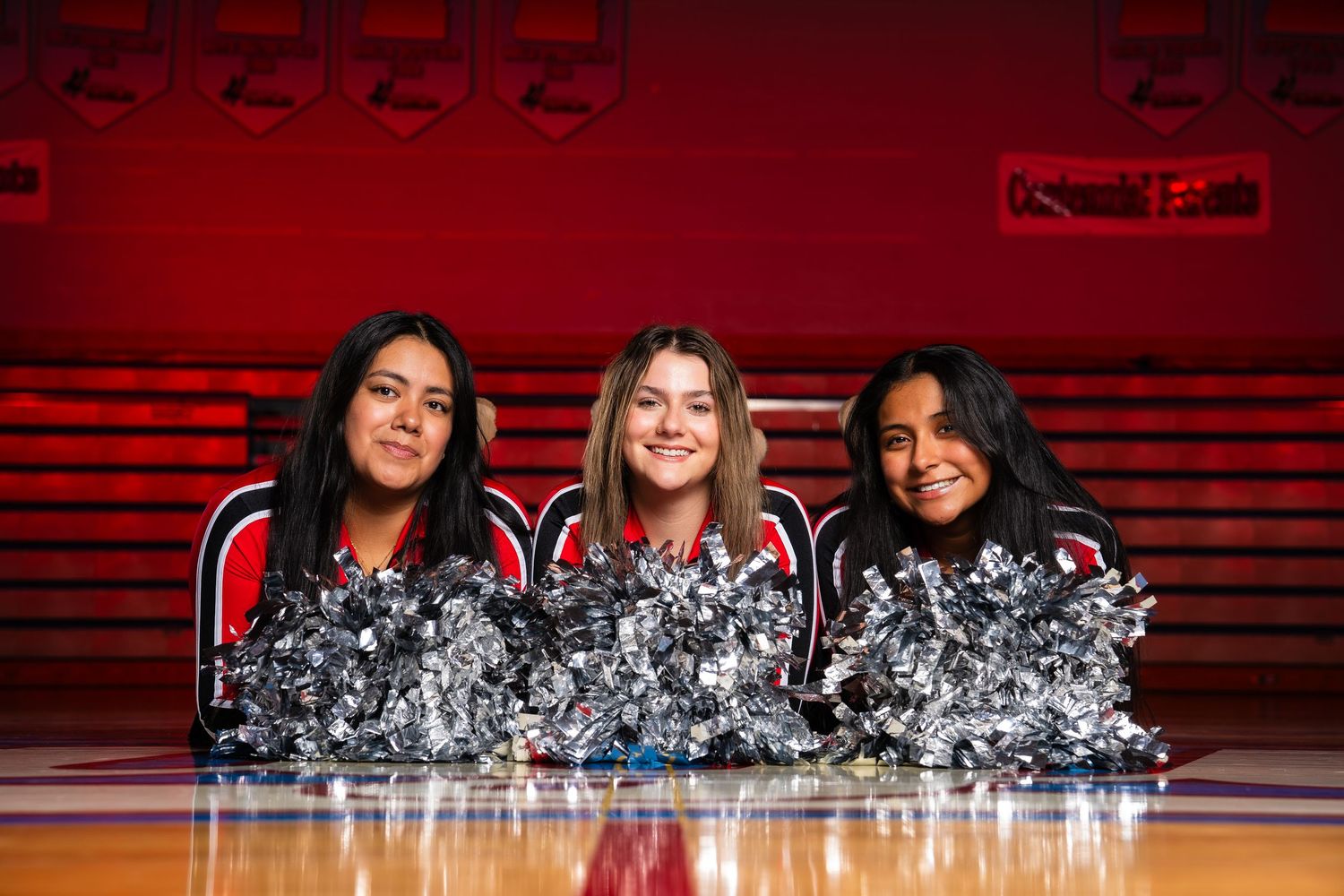 A group of cheerleaders pose together during a basketball game with red lighting and stadium seating visible in the background.