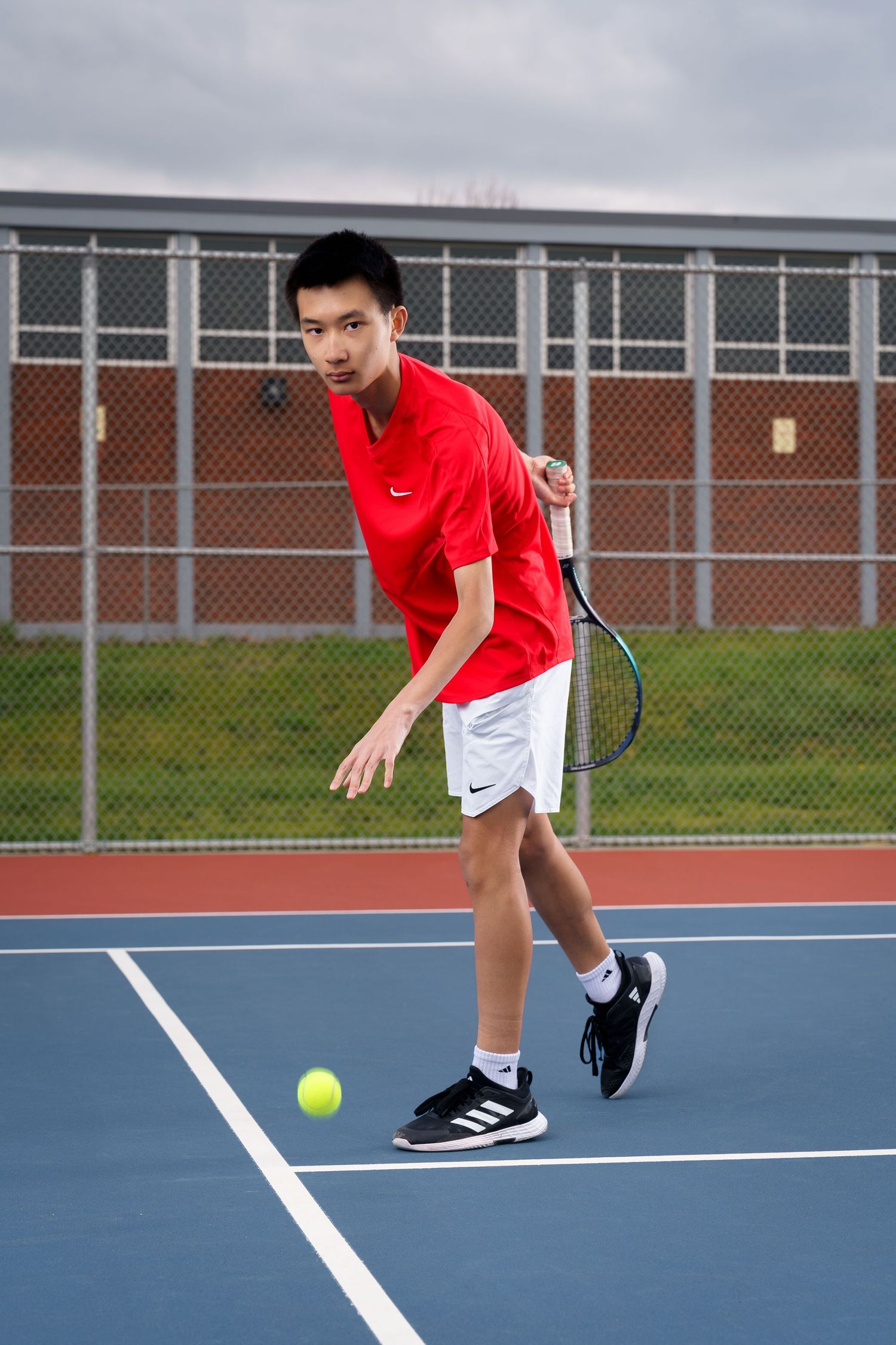 Tennis player in red shirt and black shorts reaches low to return a shot on outdoor blue court.