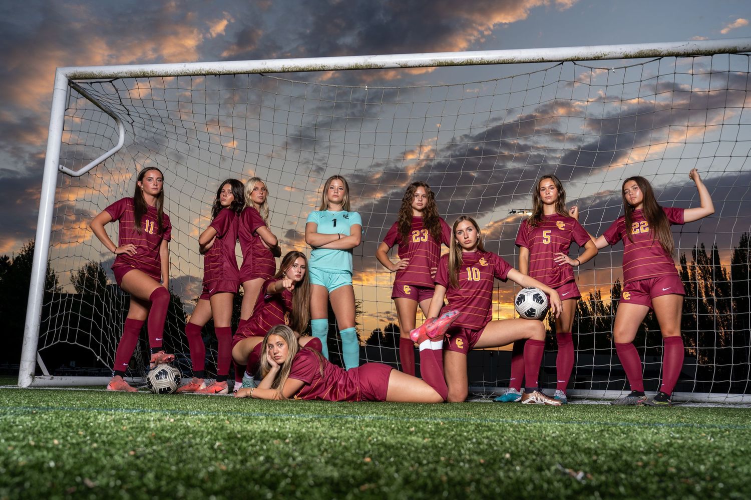 Soccer team members lay on grass field posing for creative sunset photo with goal posts in background.