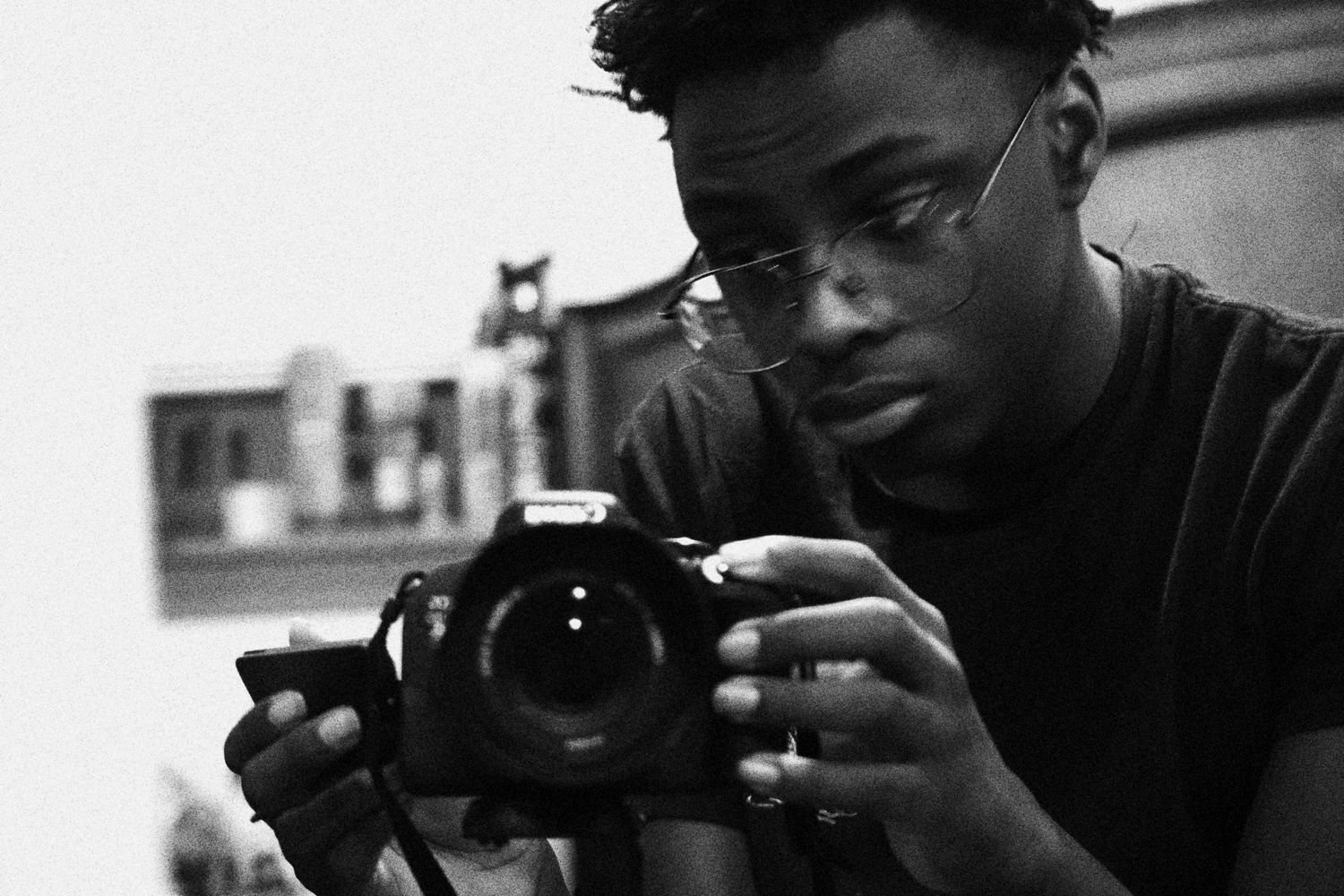 Young Black man with glasses holding a DSLR camera, captured in black and white photography.