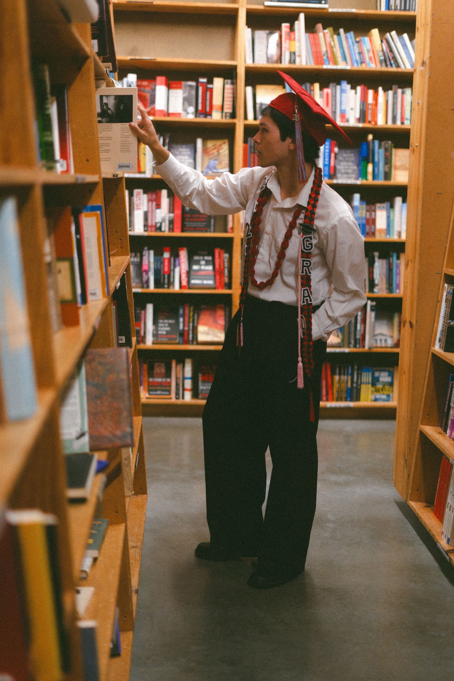 Graduate in cap and gown browsing books in a cozy bookstore aisle lined with colorful shelves.