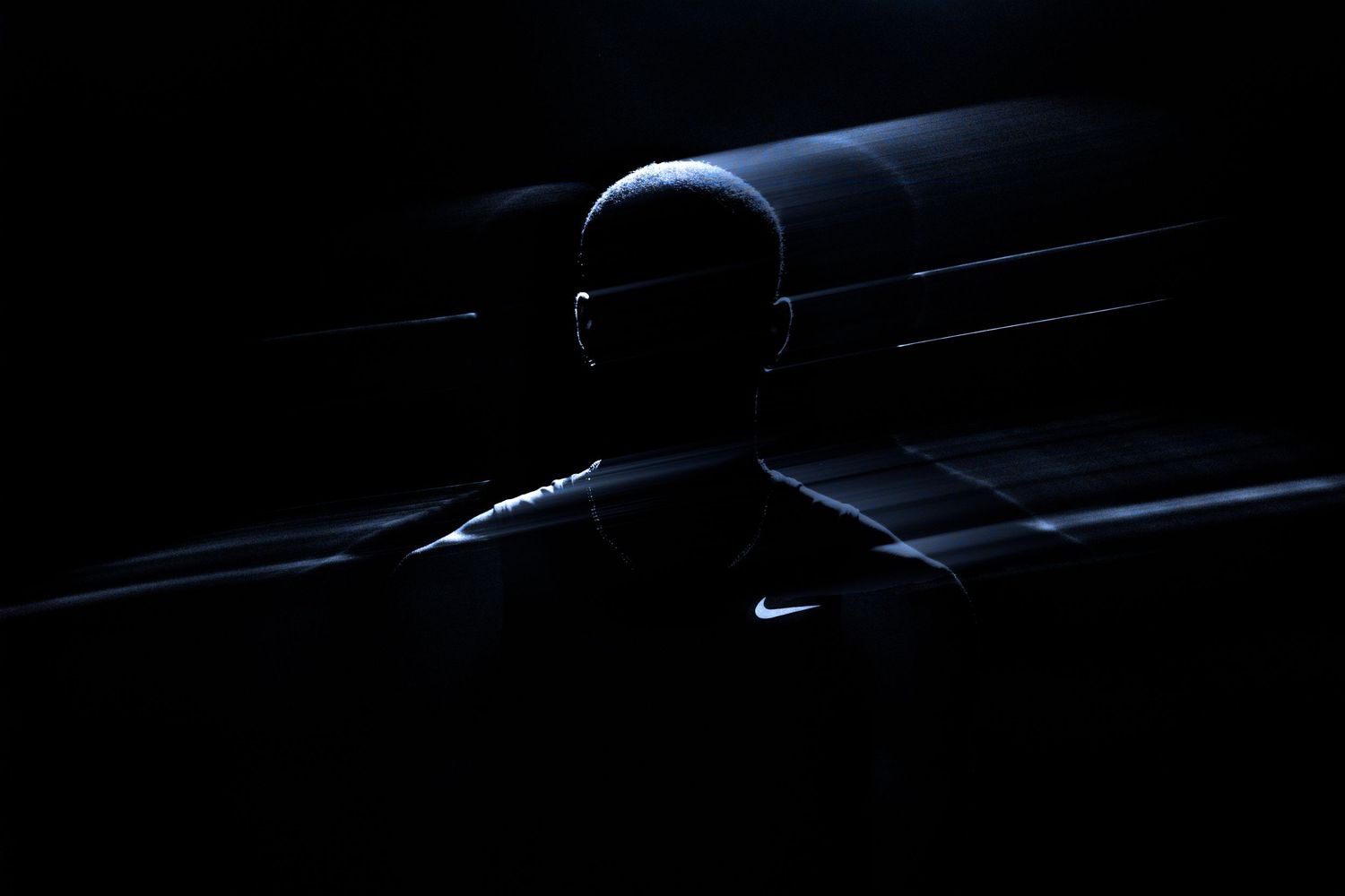 Dramatic silhouette of a bald person wearing a Nike shirt illuminated by blue light beams against a dark background.