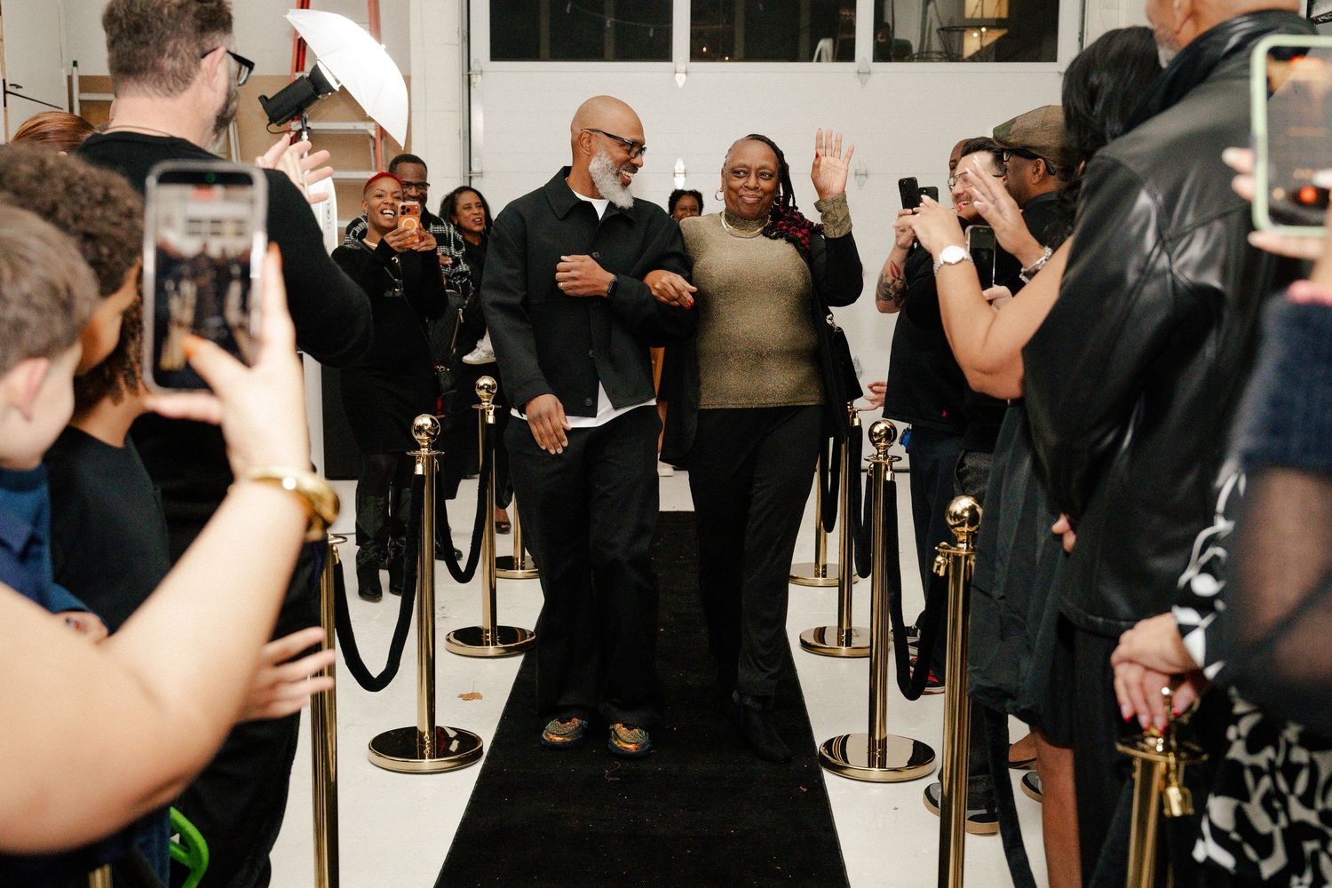Two guests walk a black carpet at a formal event, surrounded by photographers and velvet rope barriers.