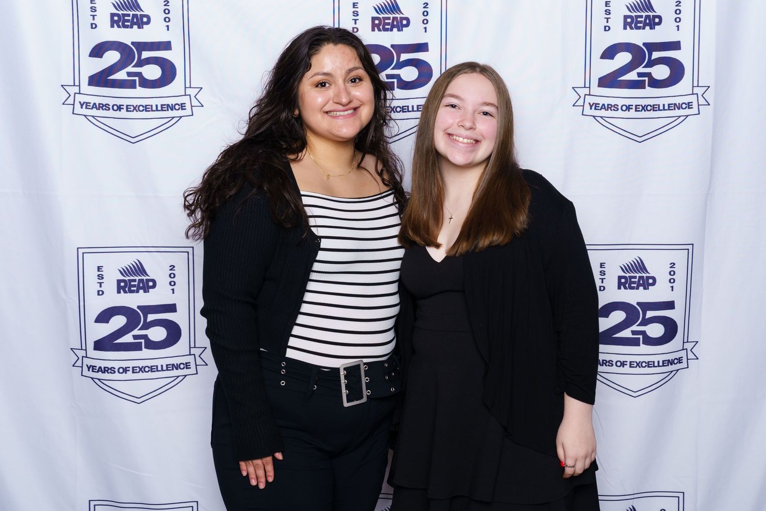 Two women smiling together in front of a REAP 25 Years of Excellence step-and-repeat banner backdrop.