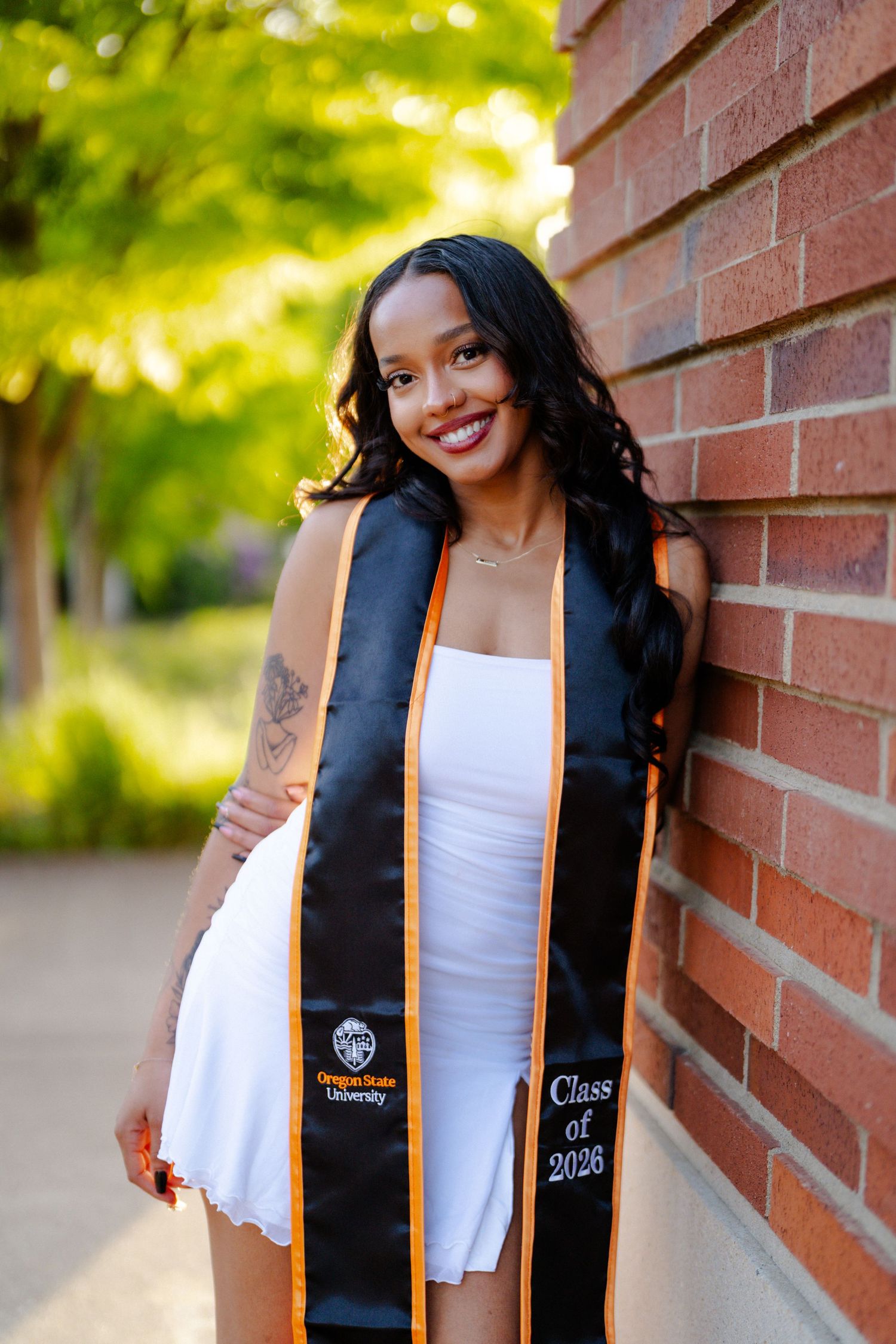 Young woman in white dress wearing Oregon State University Class of 2026 graduation stole, smiling against brick wall.