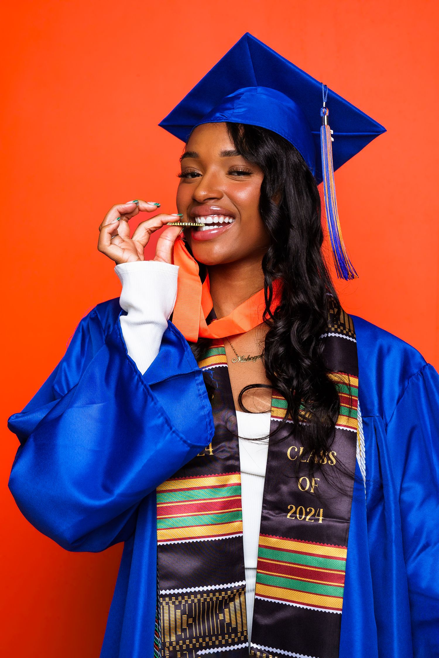 Smiling graduate in blue cap and gown with kente stole, Class of 2021, posing against a vibrant red background.