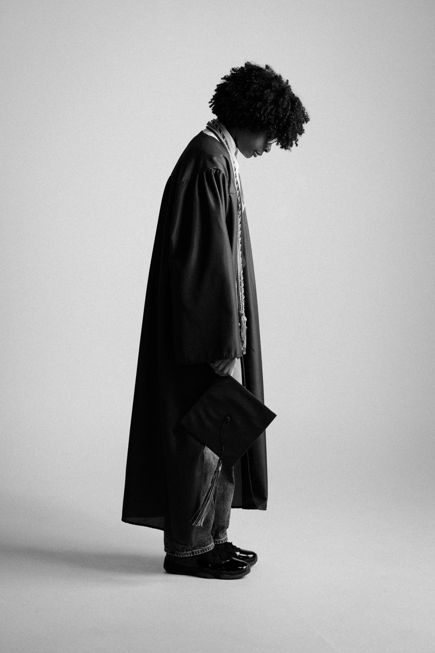 Black and white photo of a graduate in academic gown holding mortarboard, head bowed, side profile view.