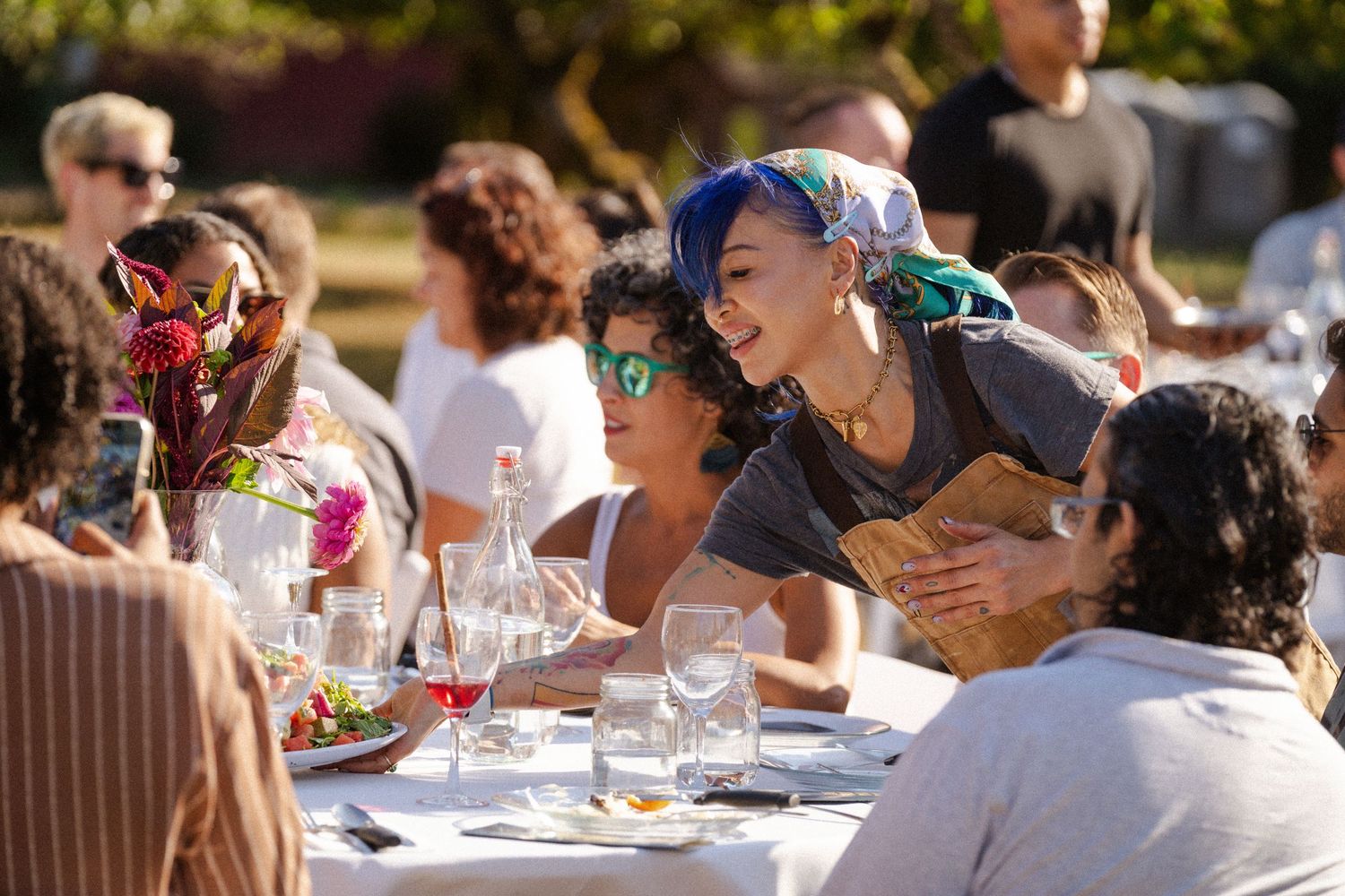 Diverse group enjoying an outdoor dinner party, with a woman in blue hair serving bread at a sunlit table.