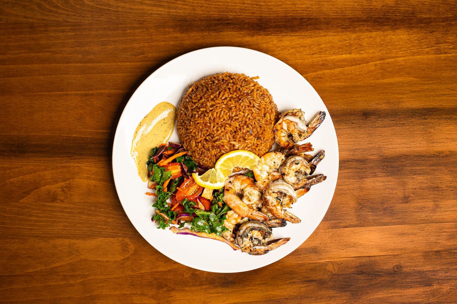 Grilled shrimp with seasoned fried rice, fresh vegetables, and lemon wedges served on a white plate.