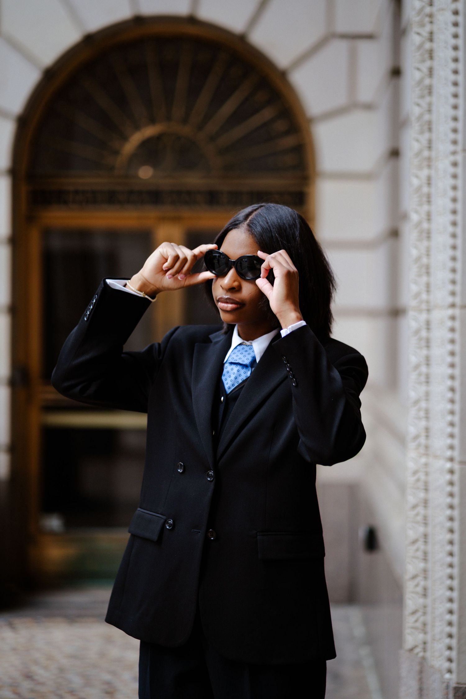 A stylish Black woman in an oversized black suit and blue tie adjusts her sunglasses outside an elegant building.