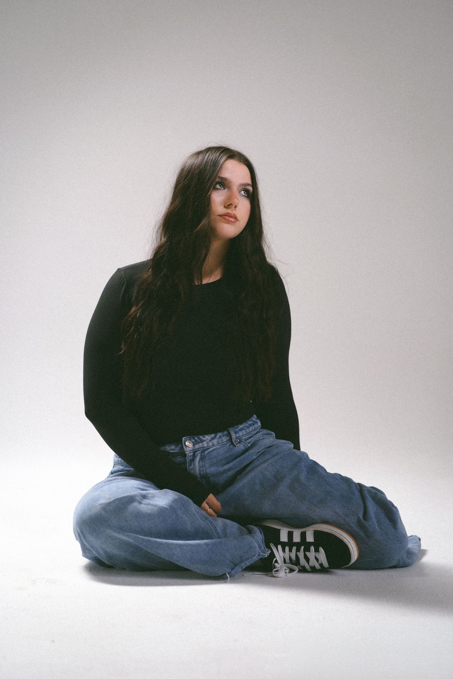 Young woman with long dark hair sits cross-legged on white floor wearing black sweater, wide-leg jeans, and dark sneakers.