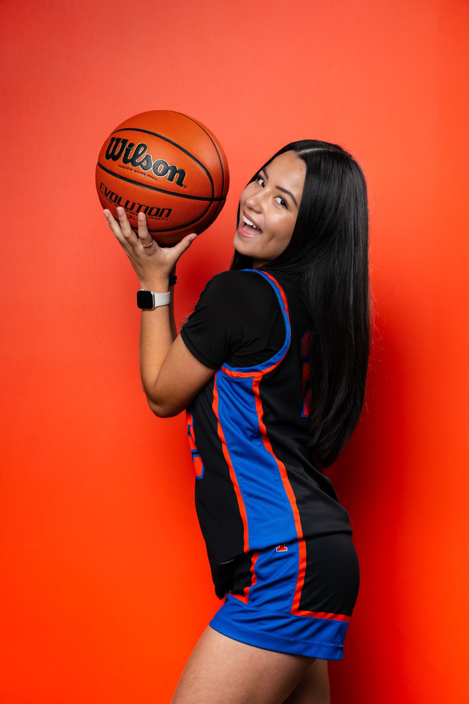 Basketball player in team jersey poses playfully with ball against vibrant red backdrop.