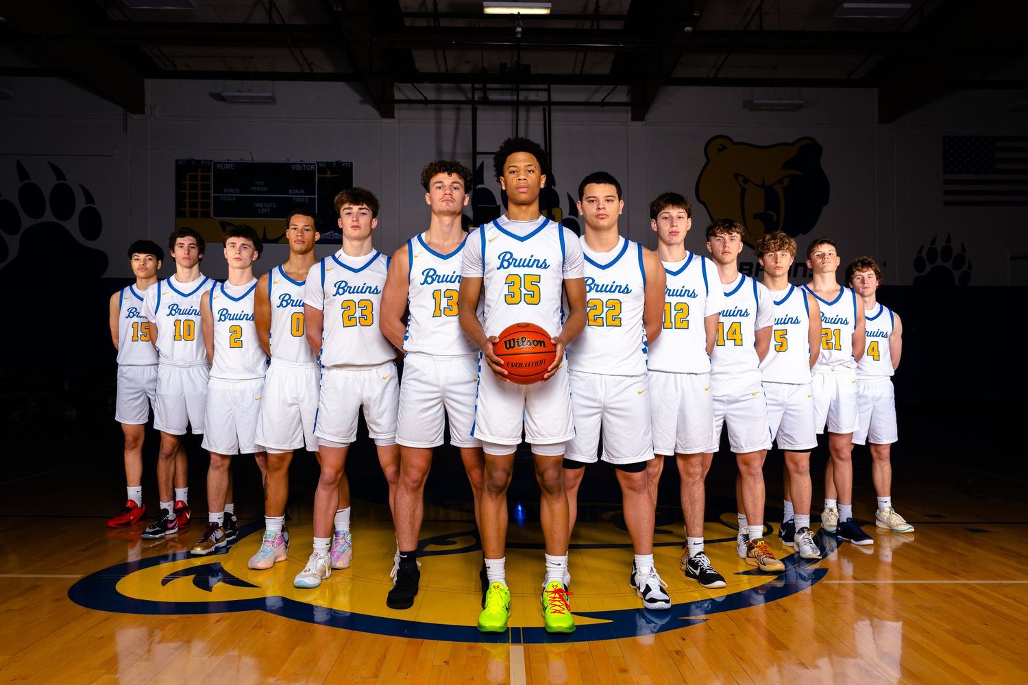 Basketball team players lined up in white uniforms on a dark court for their official team photo.