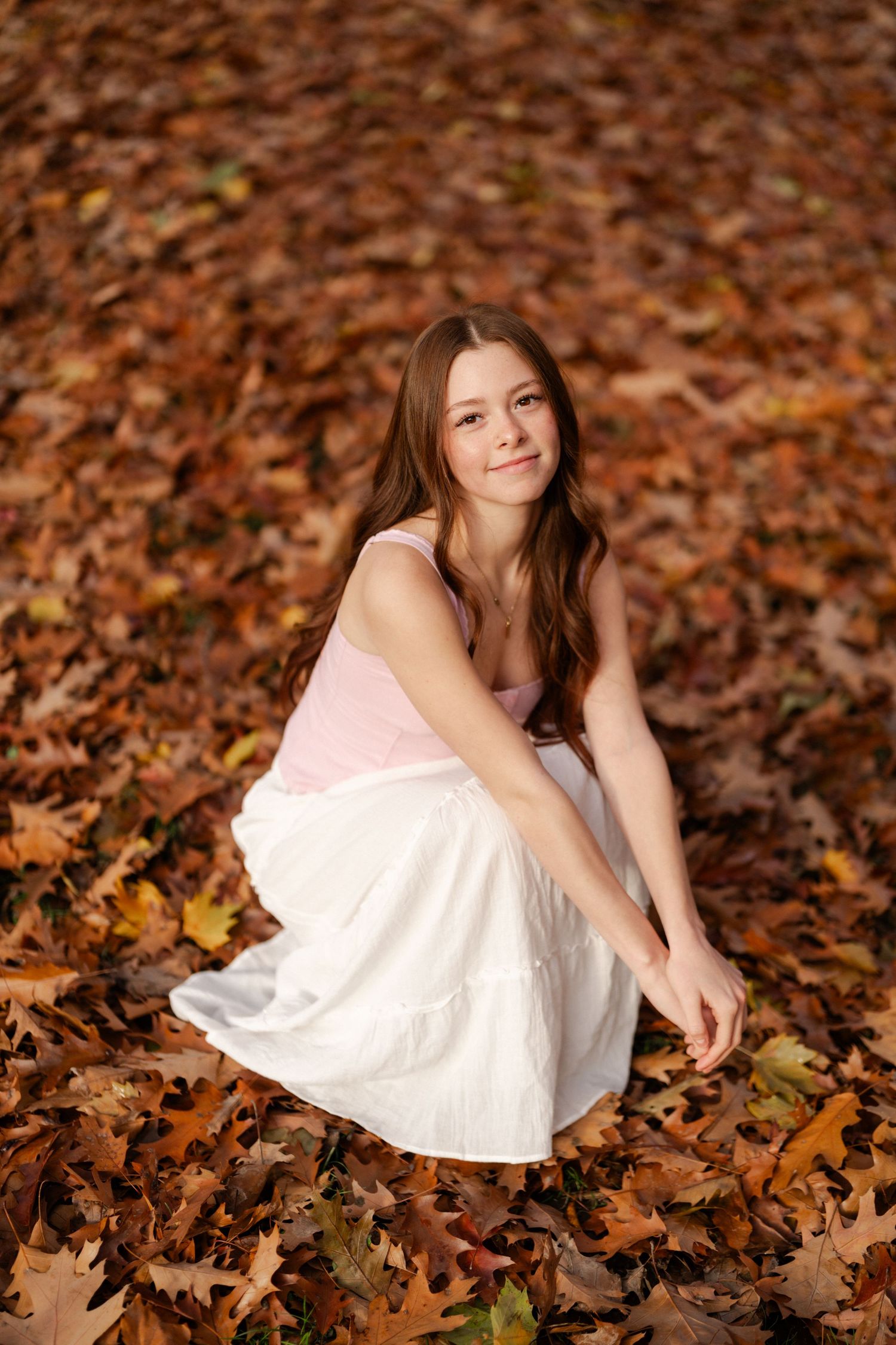 Young girl in pink top and white skirt sitting on autumn leaves in a fall forest setting.