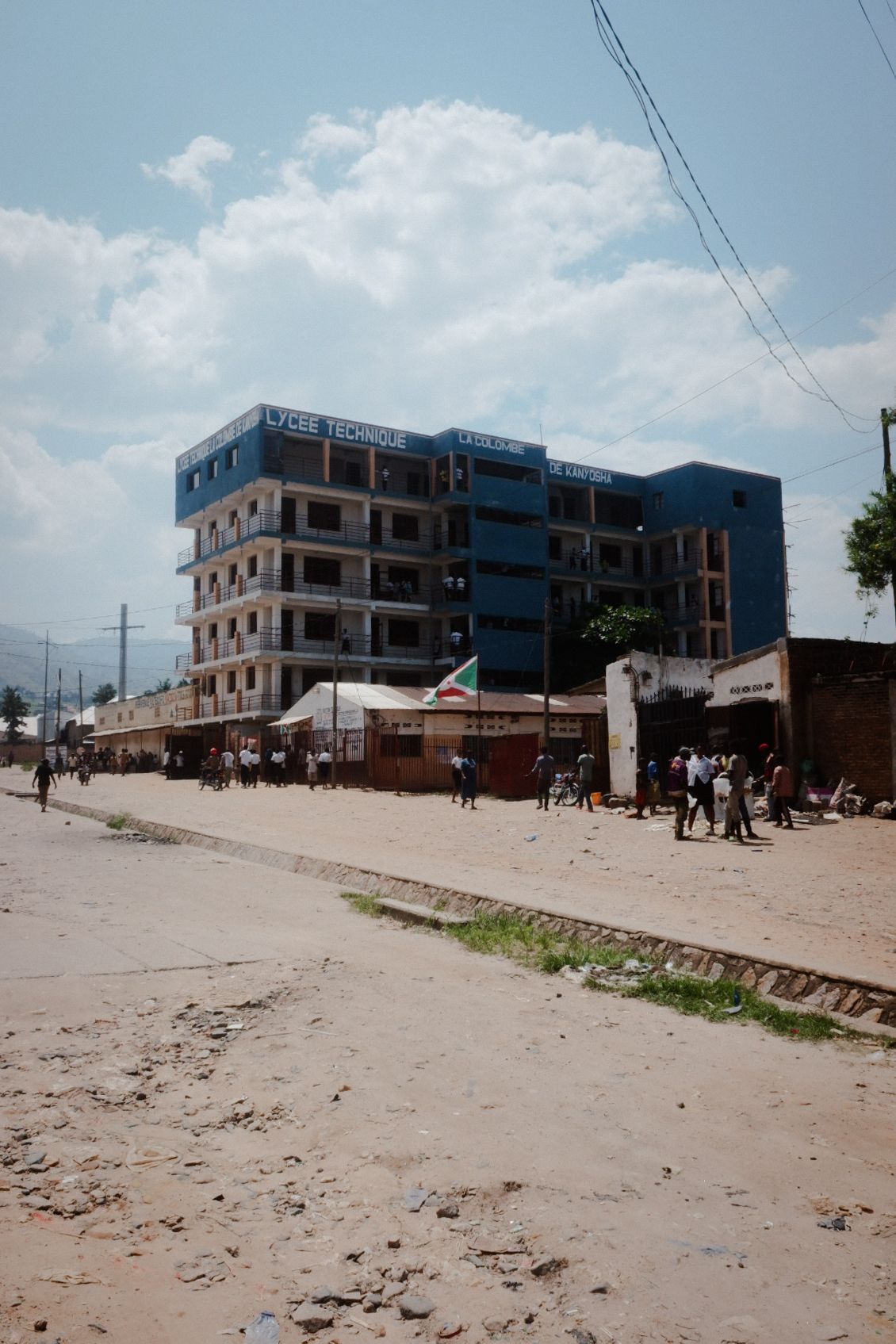 Wide dirt road with multi-story buildings and power lines in an urban area.