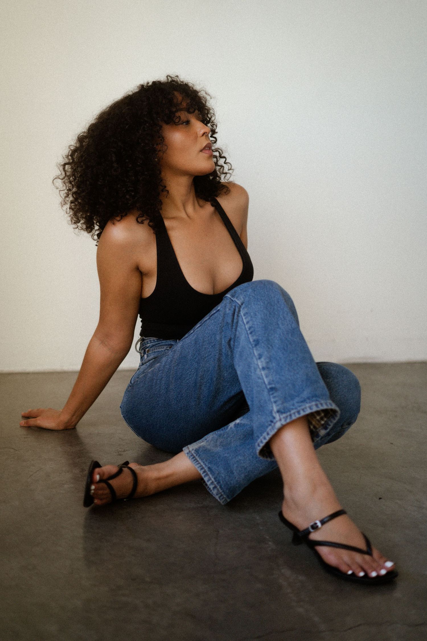 Casual pose on floor wearing jeans and sandals in minimalist setting.