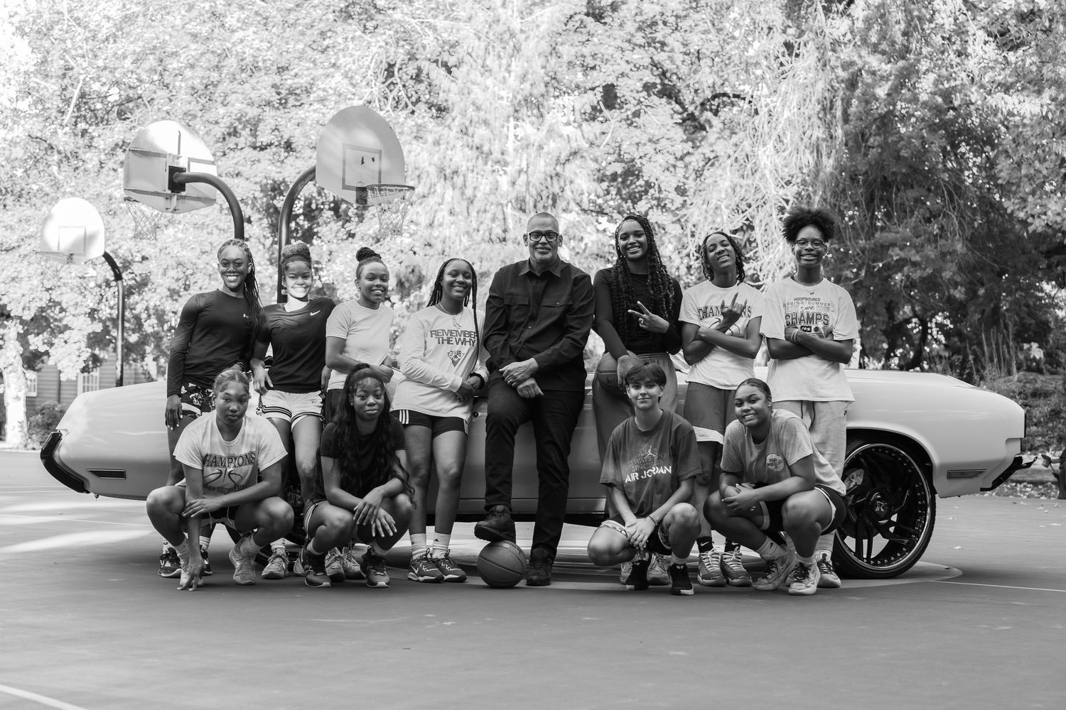 Black and white photo of a diverse group of basketball players posing together on an outdoor court near a classic car.