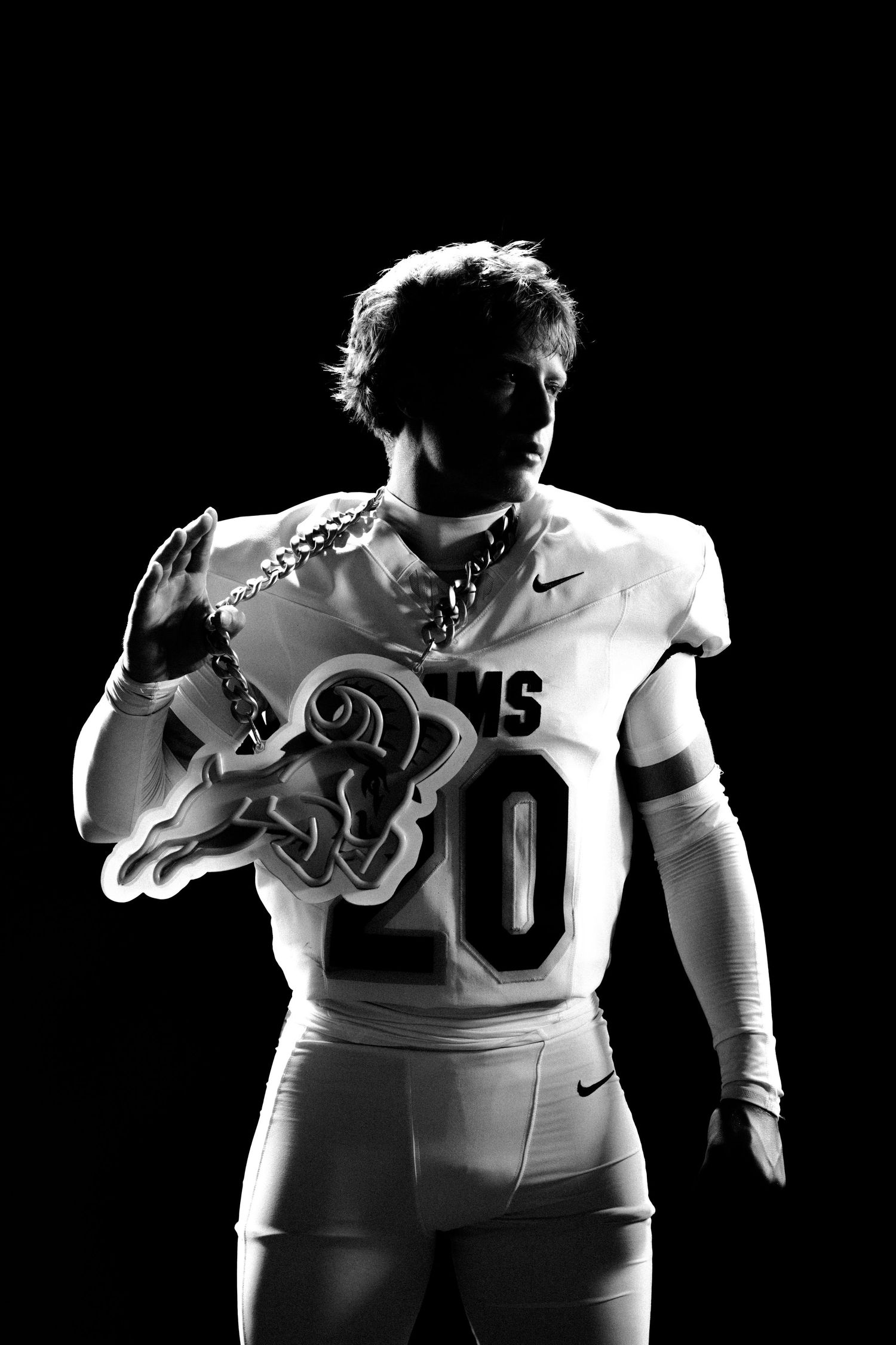 Black and white photo of a football player in white Nike jersey number 20 holding a Rams logo under dramatic lighting.