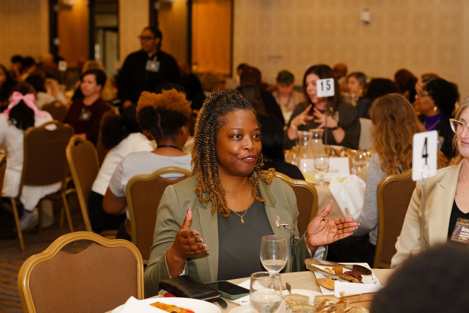 Woman gesturing expressively at a formal banquet event, seated among diverse attendees in a conference hall.