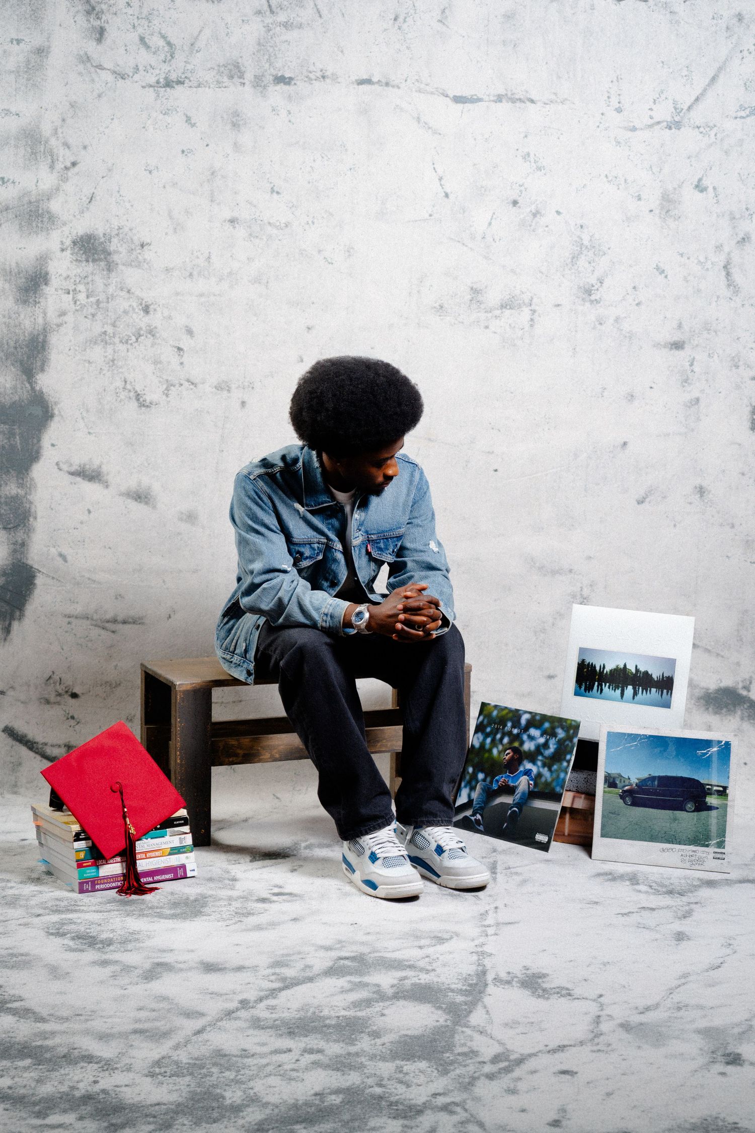 Young man in denim jacket sits on bench with graduation cap, books, and photos against concrete wall.