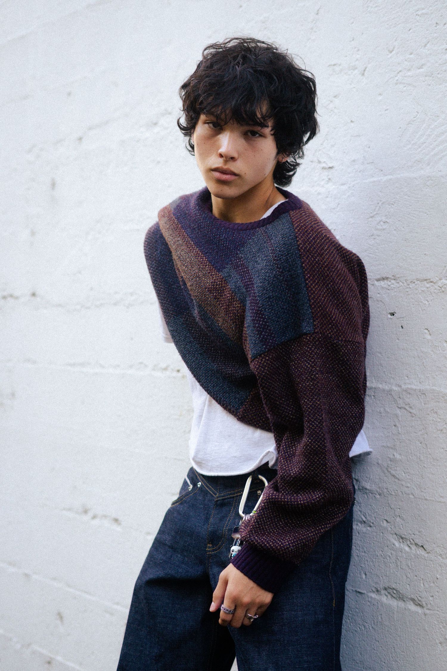 Person in purple sweater and jeans leaning against white wall in casual pose.