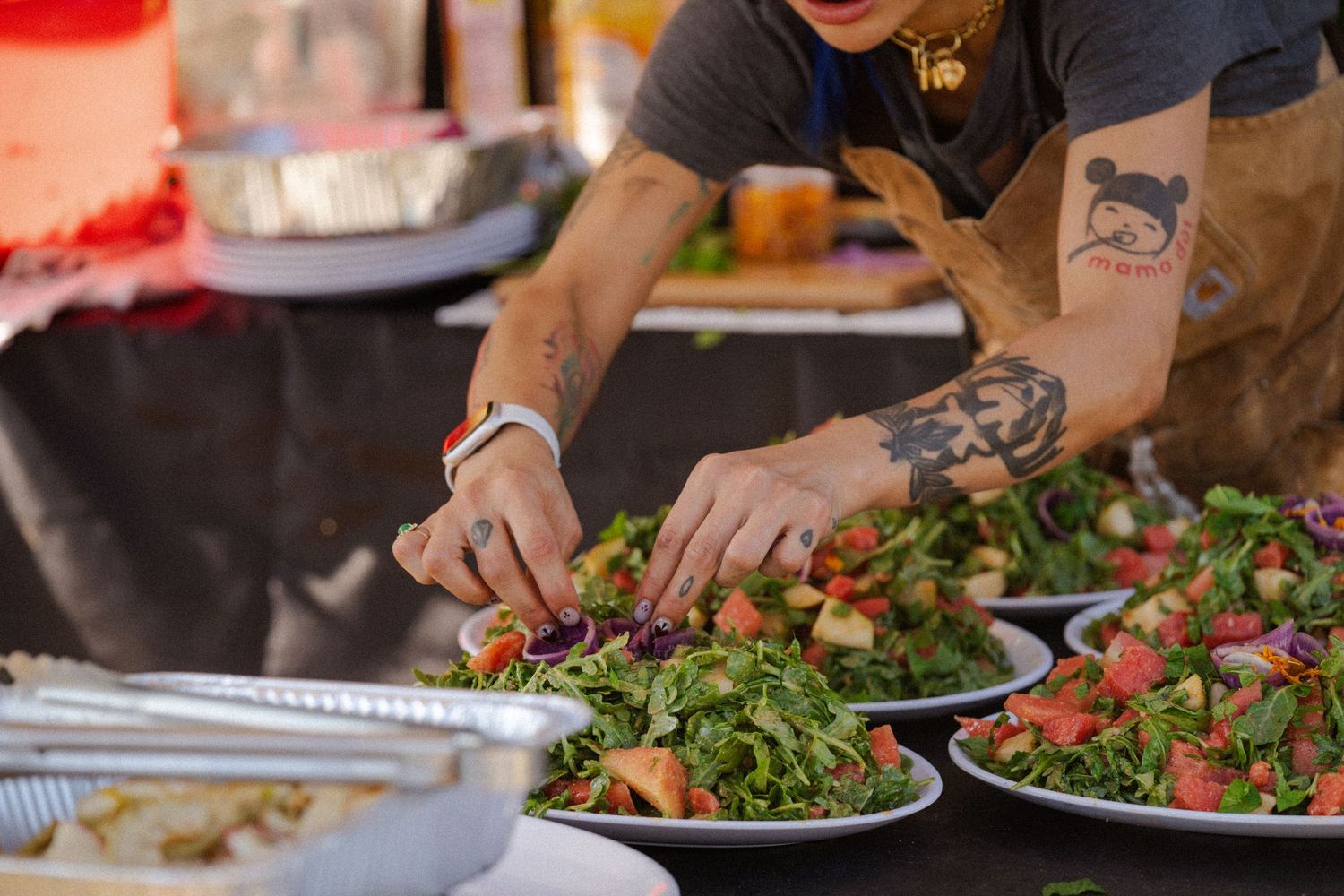 Tattooed chef arranging fresh green salads with strawberries on white plates at outdoor event.