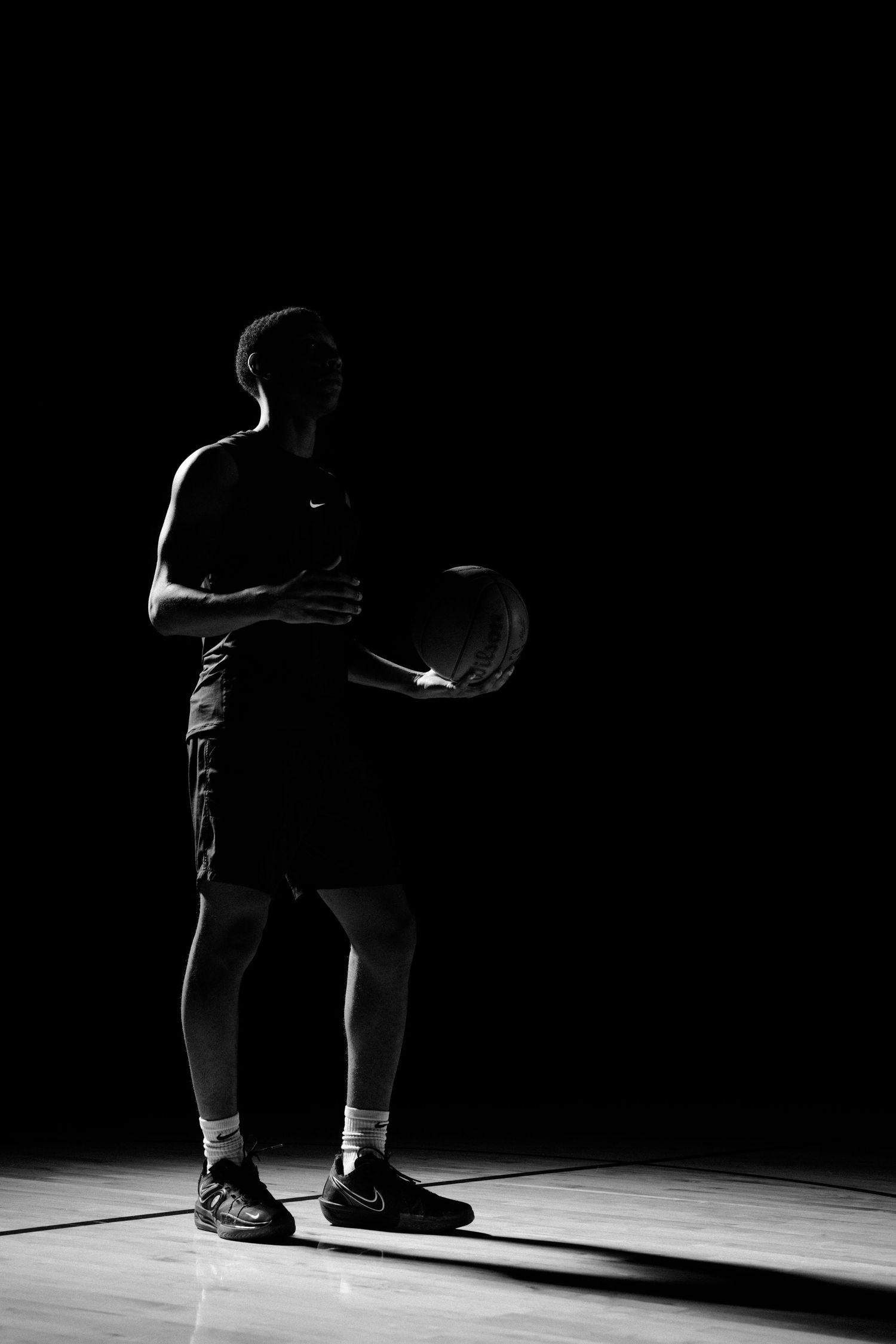 Basketball player crouches in dramatic spotlight on wooden court floor.