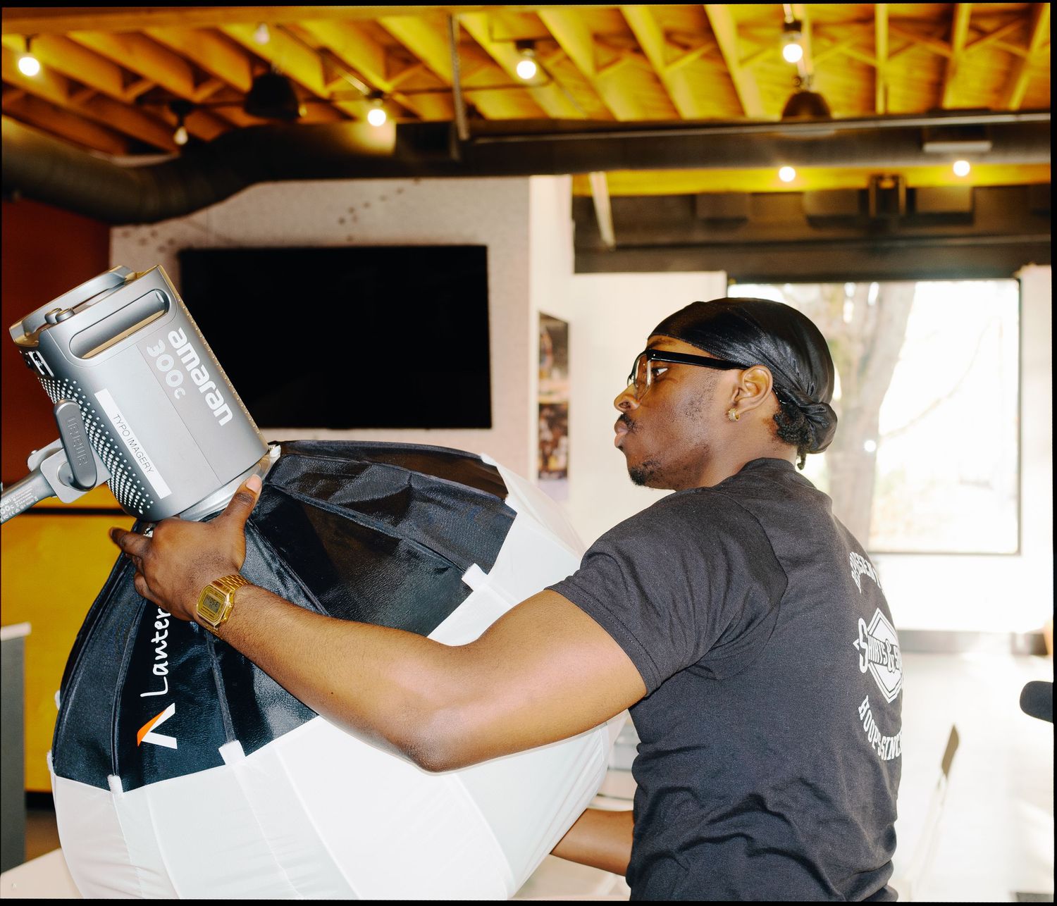 A man in a black t-shirt and sunglasses handles professional lighting equipment with a black bag in a modern interior.