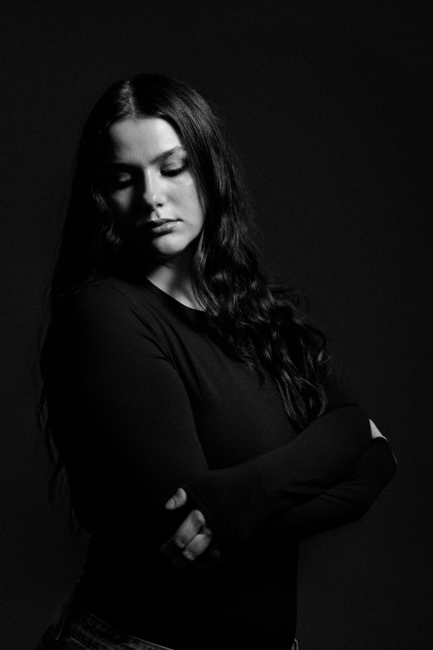 Black and white portrait of a young woman with long wavy hair, arms crossed, eyes closed, dark background.