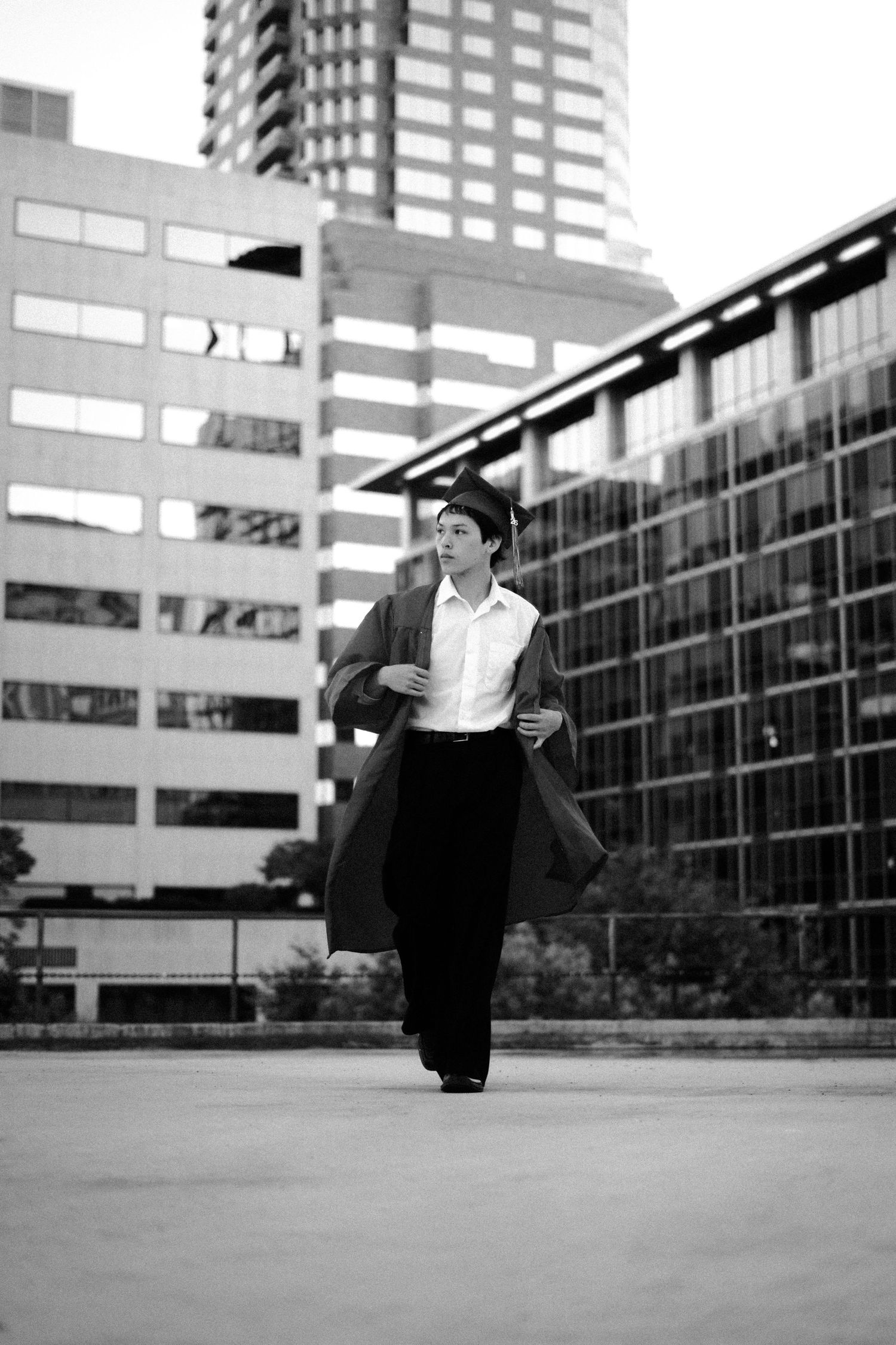 Black and white photo of a young person in graduation cap and gown walking outdoors with modern city buildings in background.