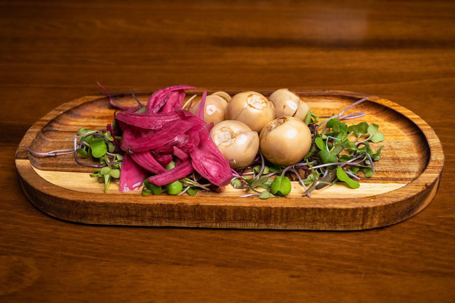 Wooden board with pickled red onions, whole mushrooms, and fresh microgreens on a table.