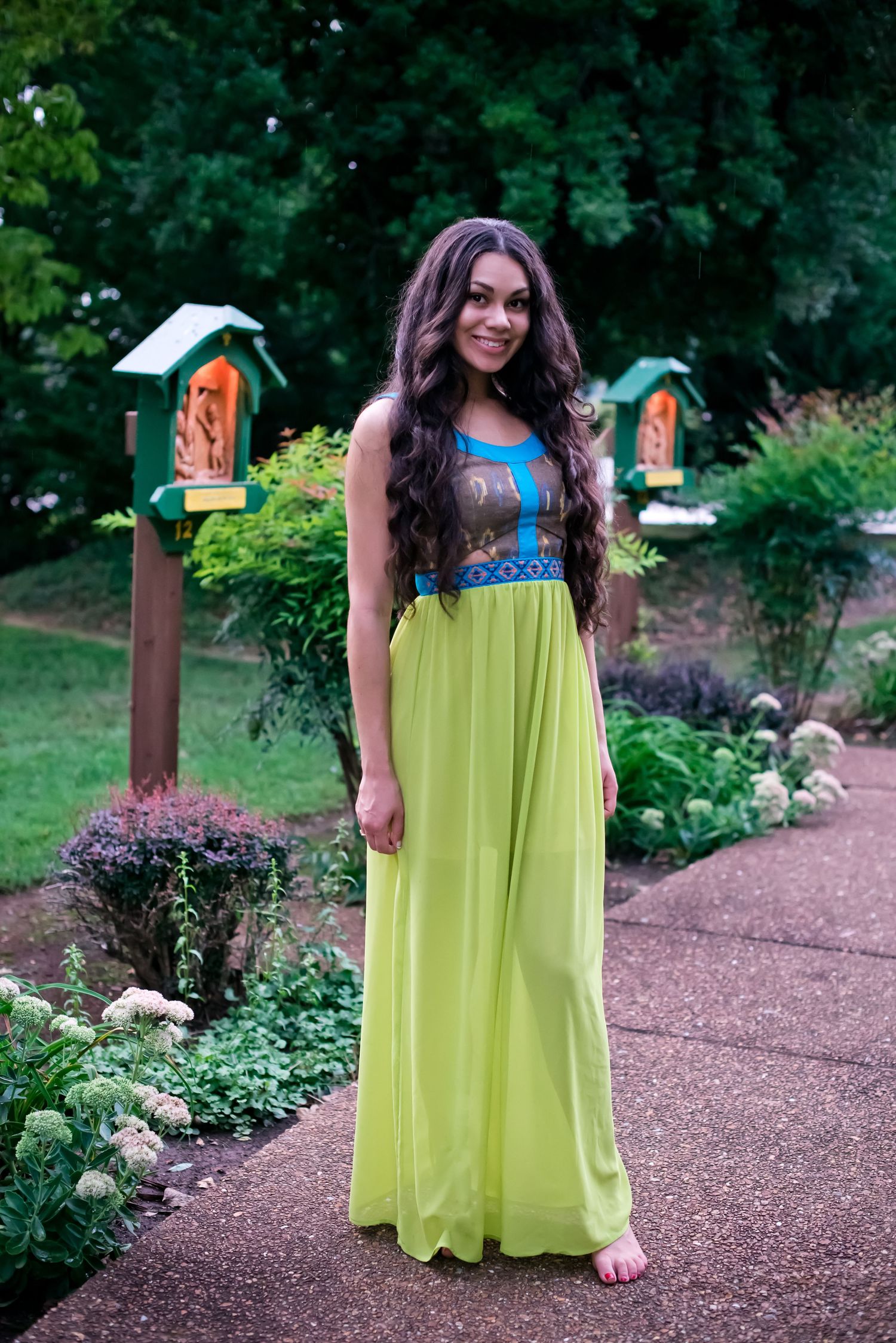 A person wearing a bright yellow-green flowing dress poses on a garden path surrounded by lush greenery and flowers.