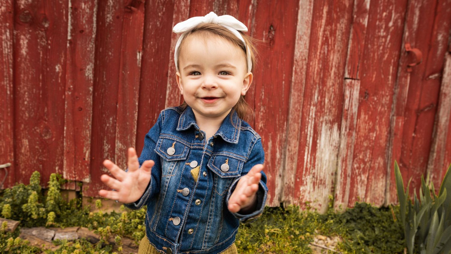 Toddler in denim jacket smiling against red barn wall background.