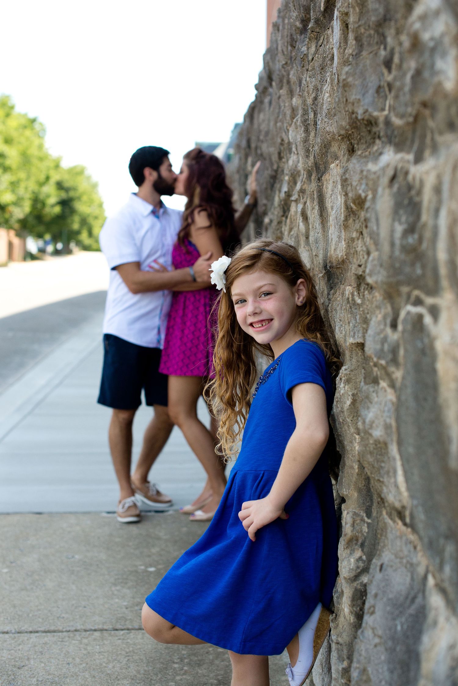 A young girl in a blue dress poses near a stone wall while a couple embraces in the background.