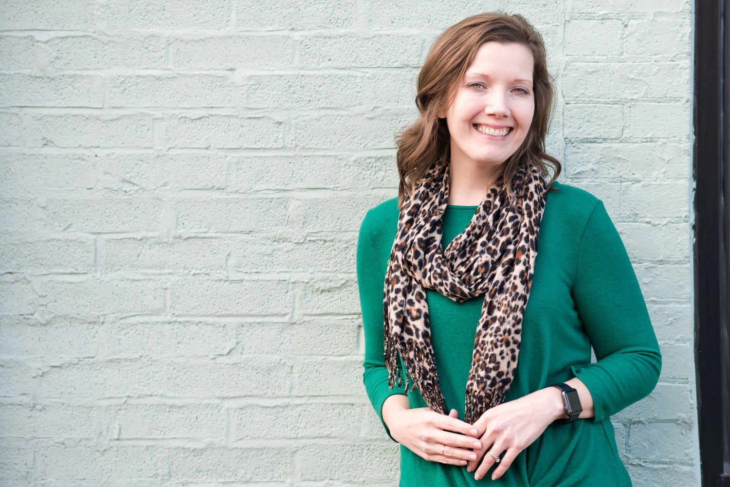 Woman in green sweater and leopard print scarf against white brick wall.