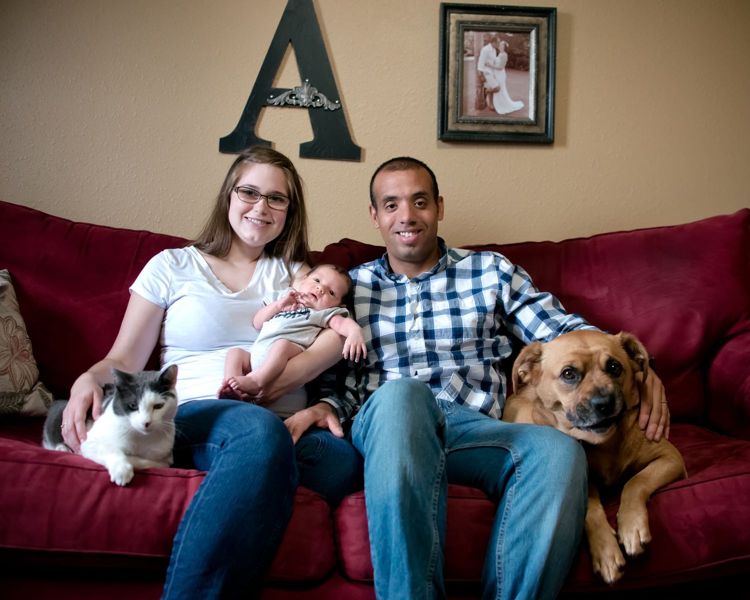 Family sitting on red couch at home with their pets including a cat and dog, with decorative letter A on wall.