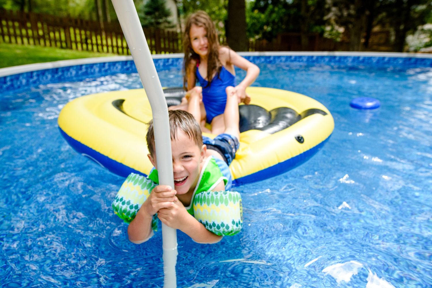 Children playing and swimming in backyard pool with yellow float on sunny summer day.