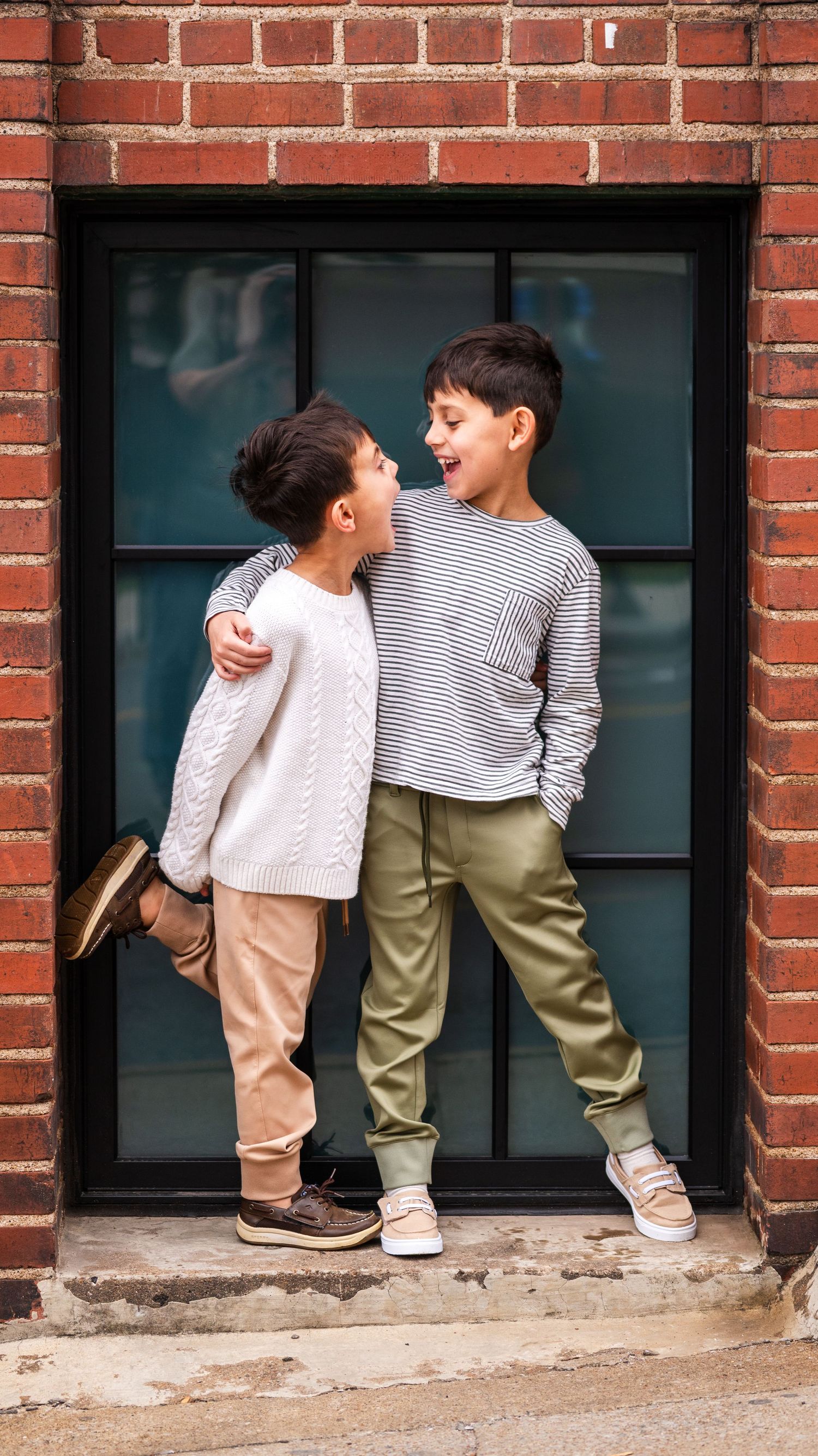 Two children in casual wear laughing together in doorway against brick wall.