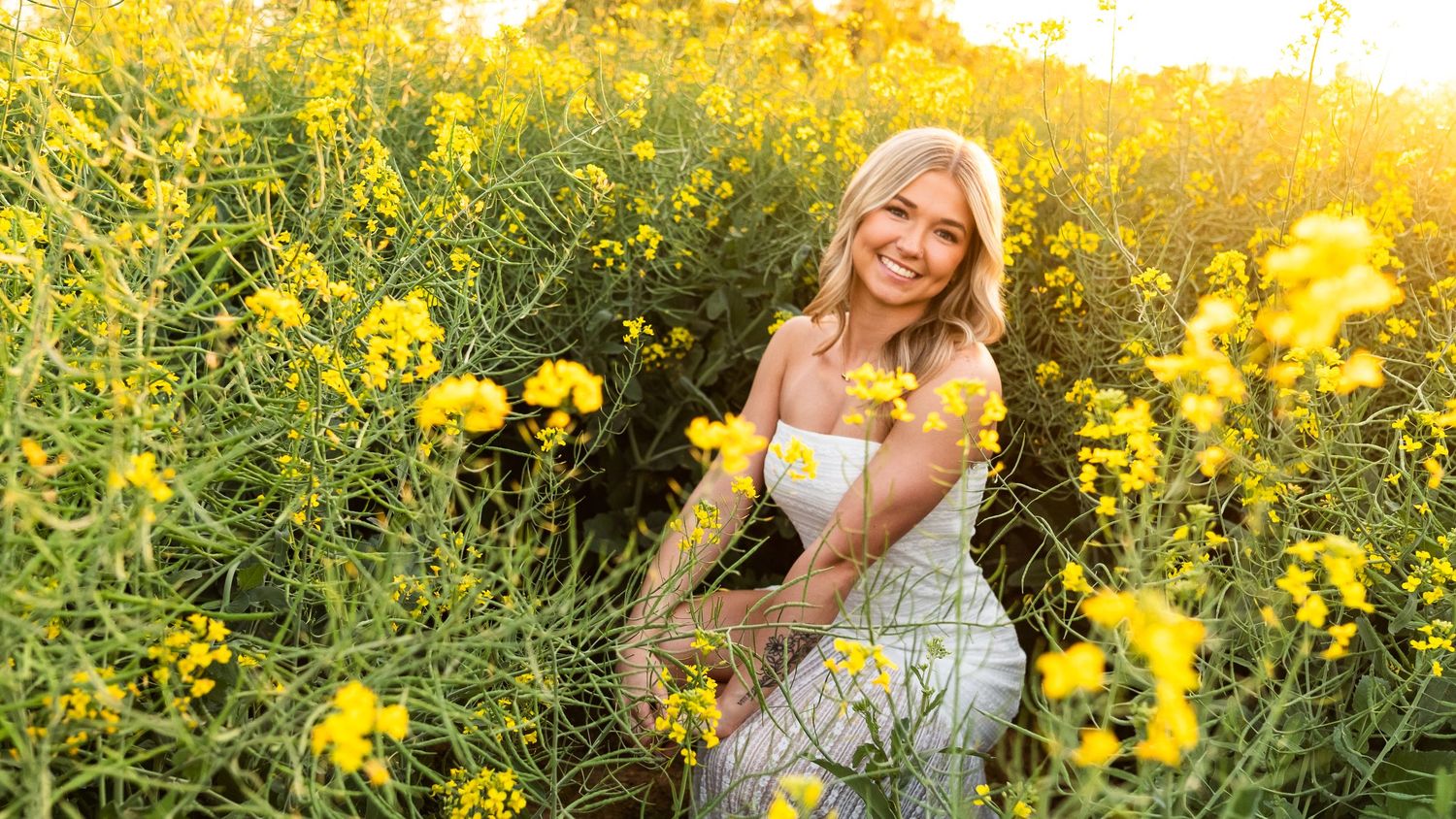 Smiling blonde woman in white dress surrounded by vibrant yellow wildflowers in a sunny field.